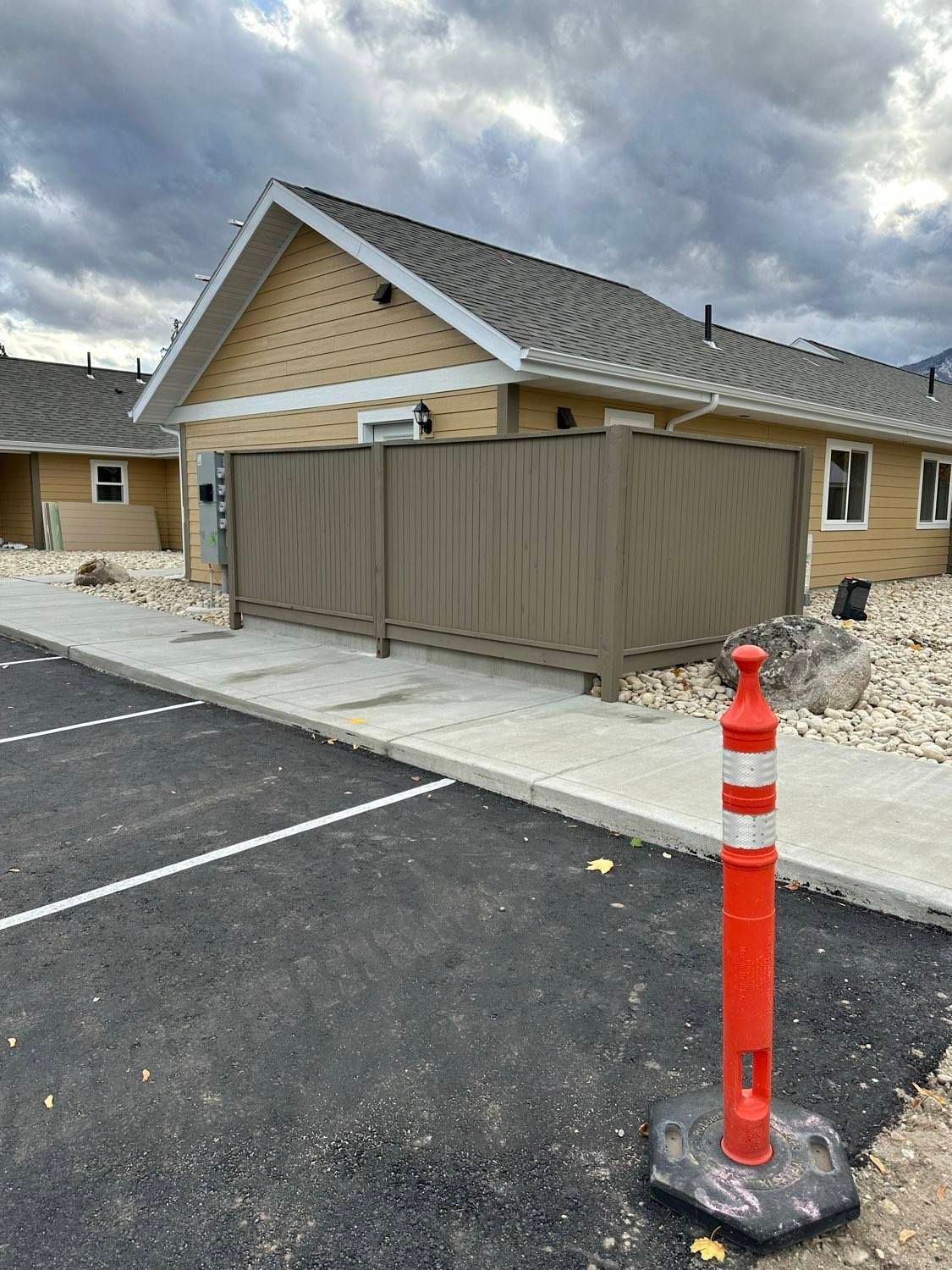 Tan building with brown fence, concrete sidewalk, and red and white bollard. Black asphalt parking lot.