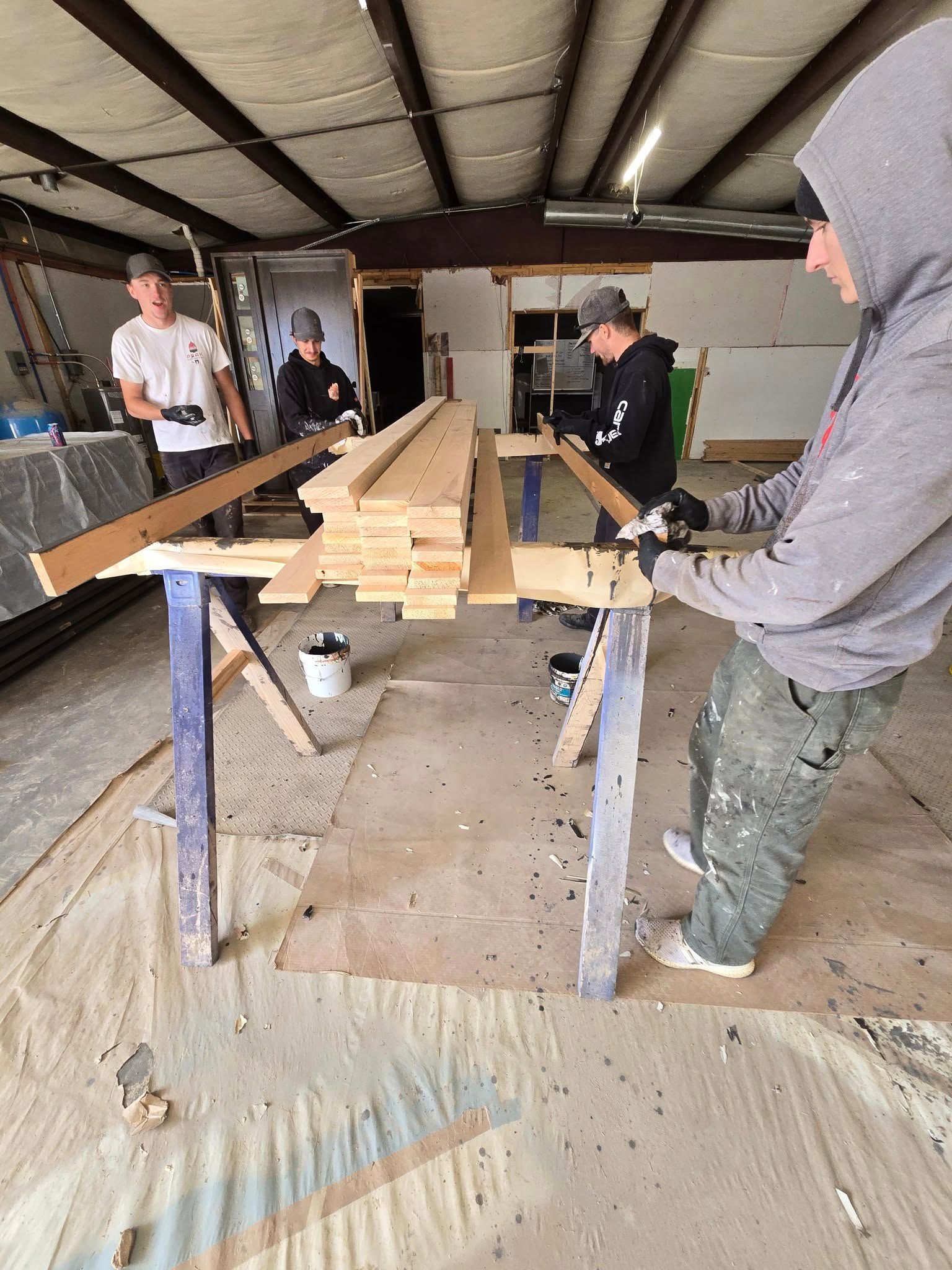 Four people working with wood on a workbench inside a workshop. One holds wood, others paint.