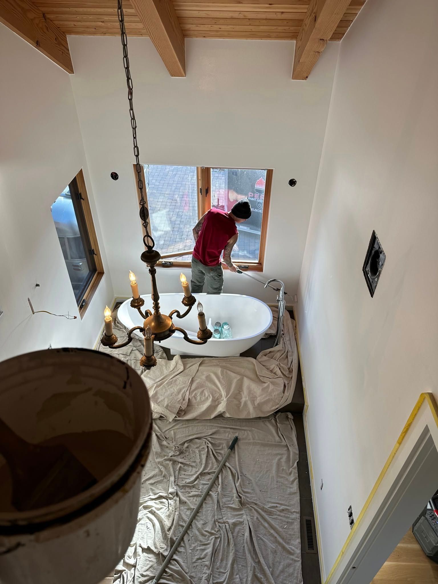 Person painting around a window in a bathroom with a large tub and chandelier.
