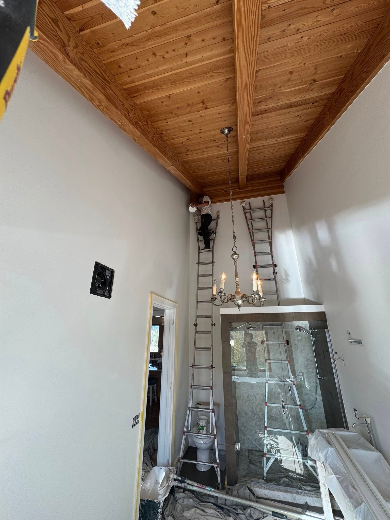 Person on tall ladder near chandelier in entryway; unfinished ceiling with wood beams, white walls.