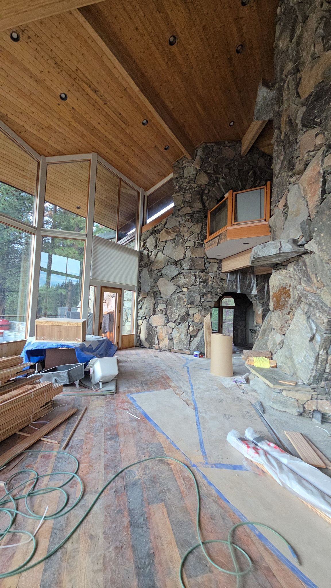Interior of a home under construction with a large stone fireplace, high wood ceilings, and large windows.