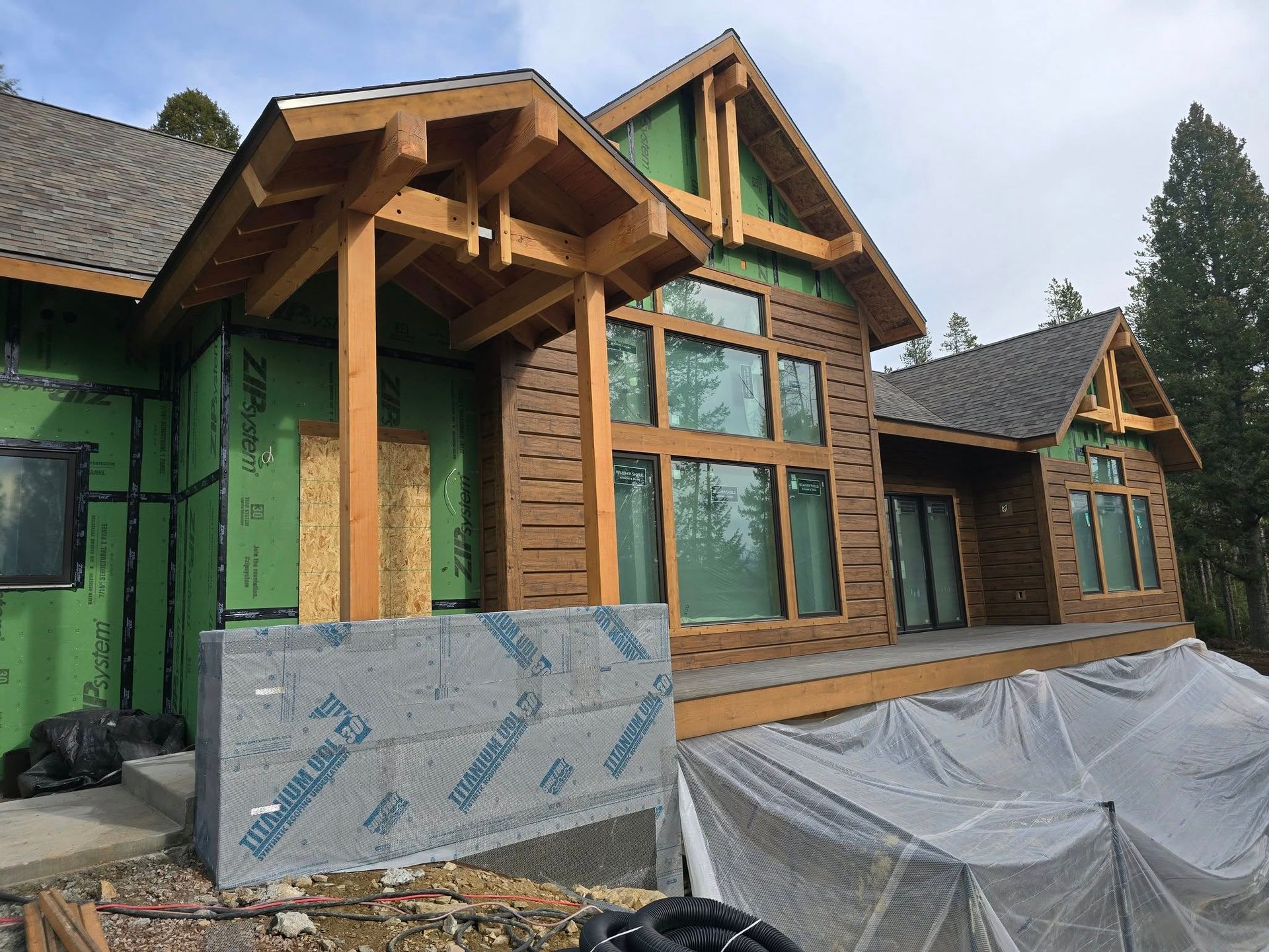 House under construction with brown siding, wooden porch, green insulation, and large windows.