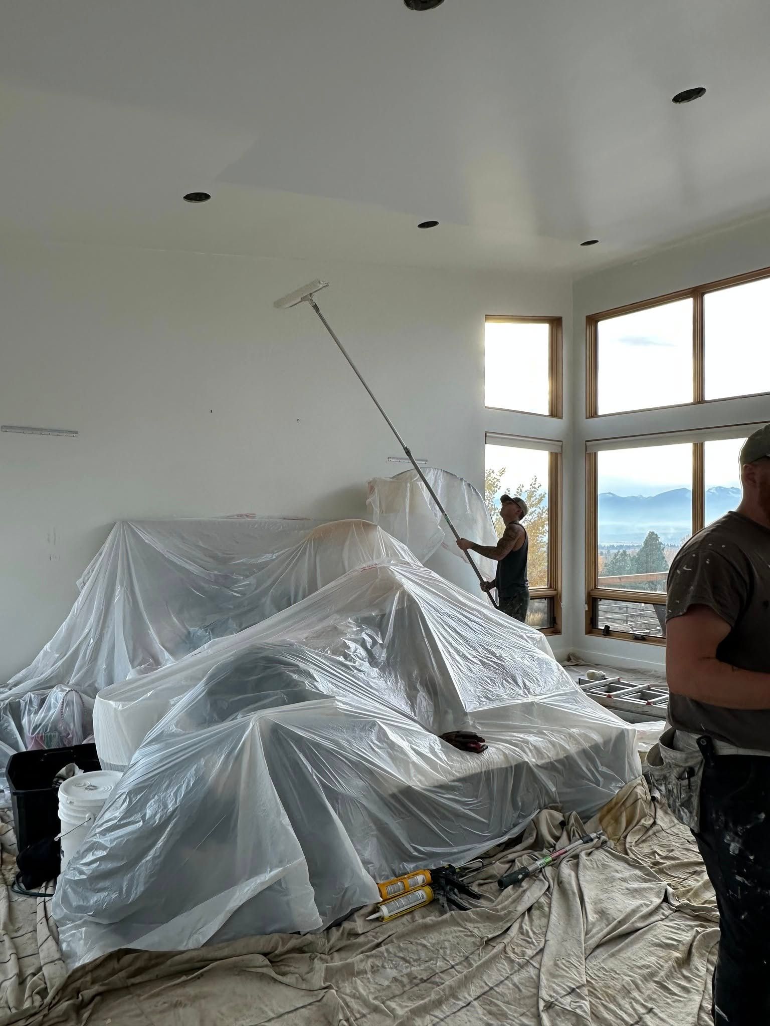 Two people painting a white interior room. One uses a roller on the ceiling, while furniture is covered in plastic sheeting.