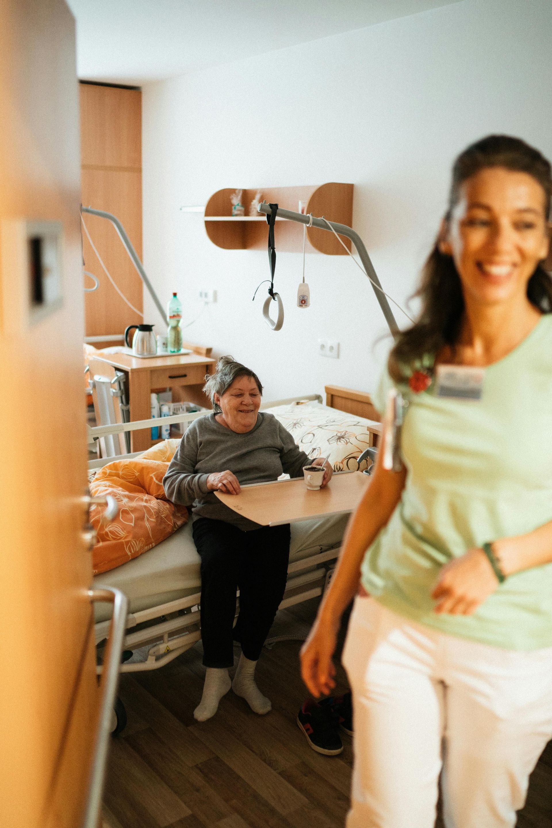 A smiling caregiver stands by a bed with a resident in a care facility.