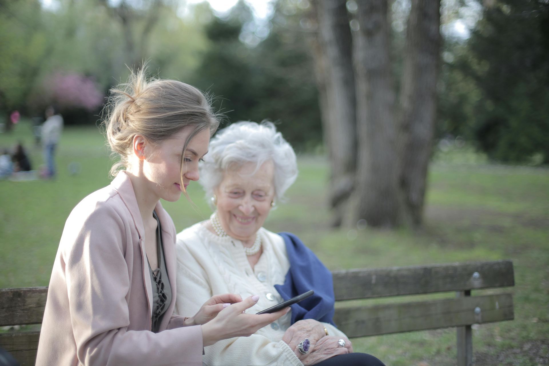 Young woman showing a cell phone to an elderly woman seated on a park bench. They are smiling.