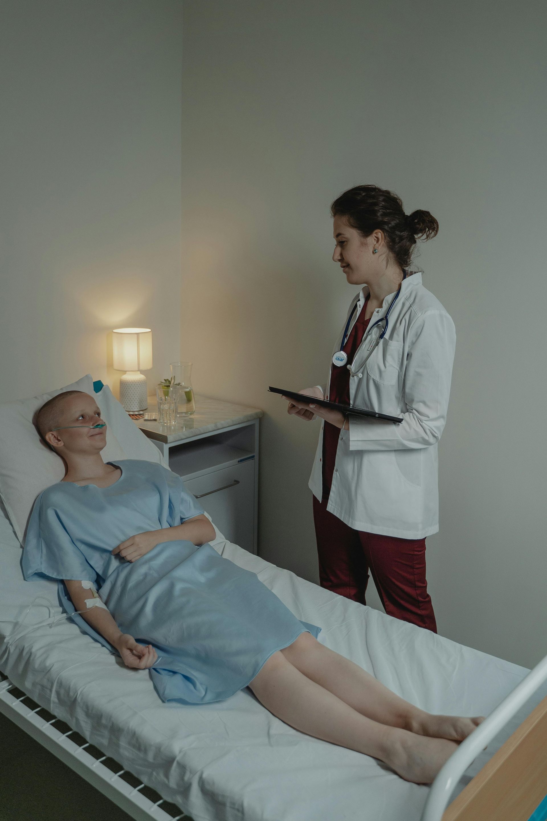 Doctor speaks with patient in bed. Patient in gown, doctor holding tablet. White room with bedside table.