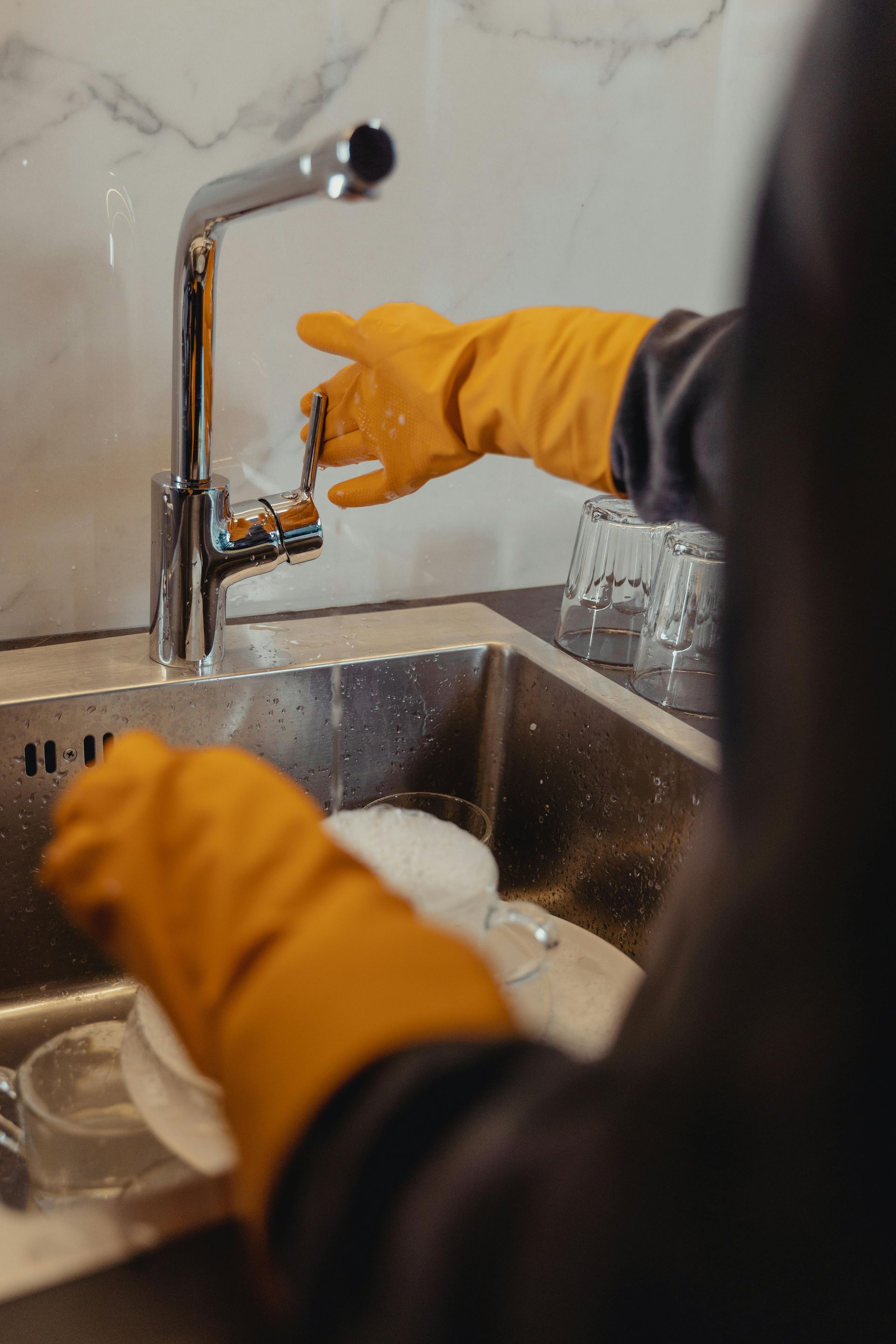 Person wearing orange gloves washing dishes in a stainless steel sink with a chrome faucet.