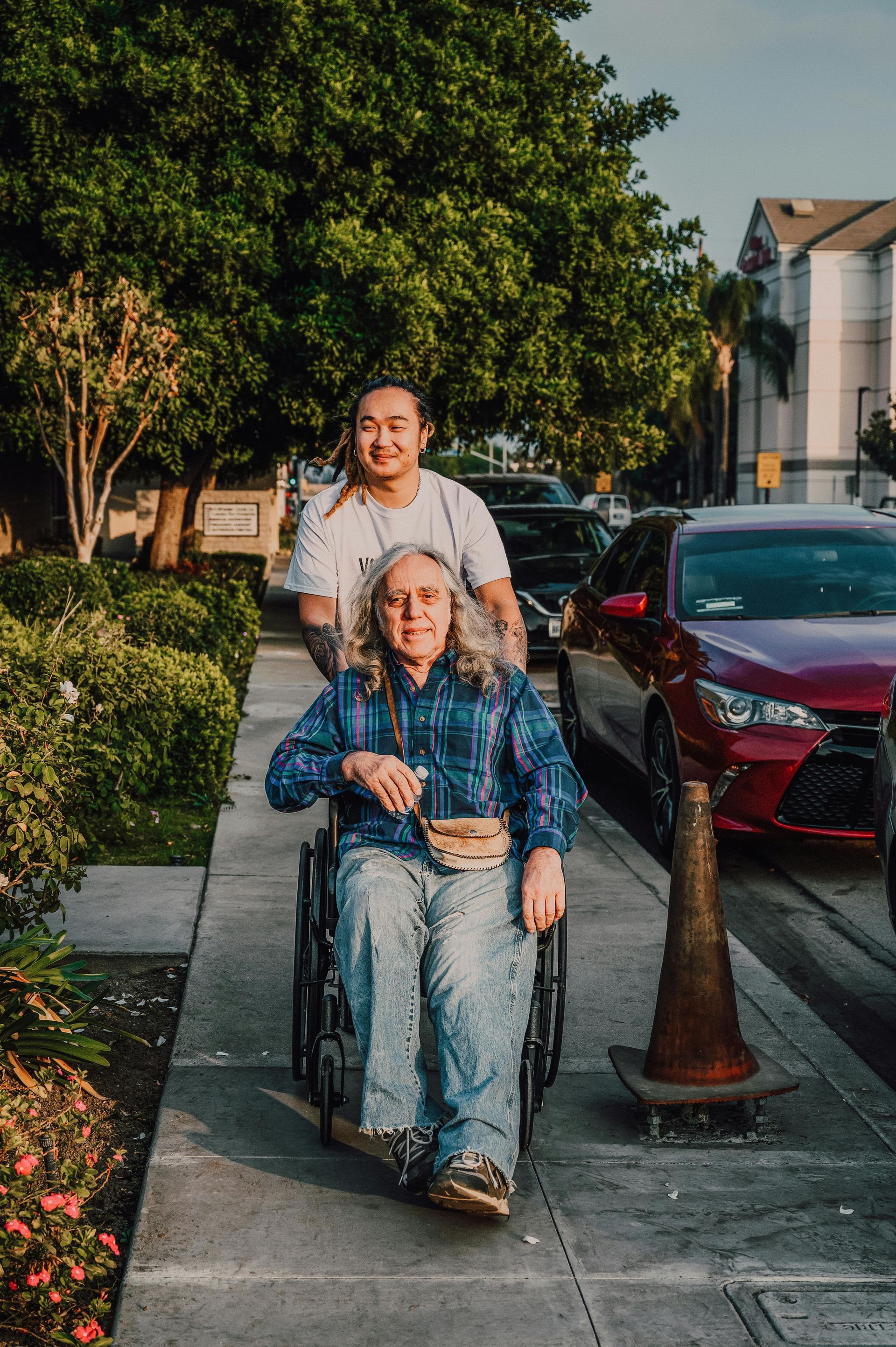 Person pushing a person in a wheelchair on a sidewalk next to parked cars. Trees and a building in background.