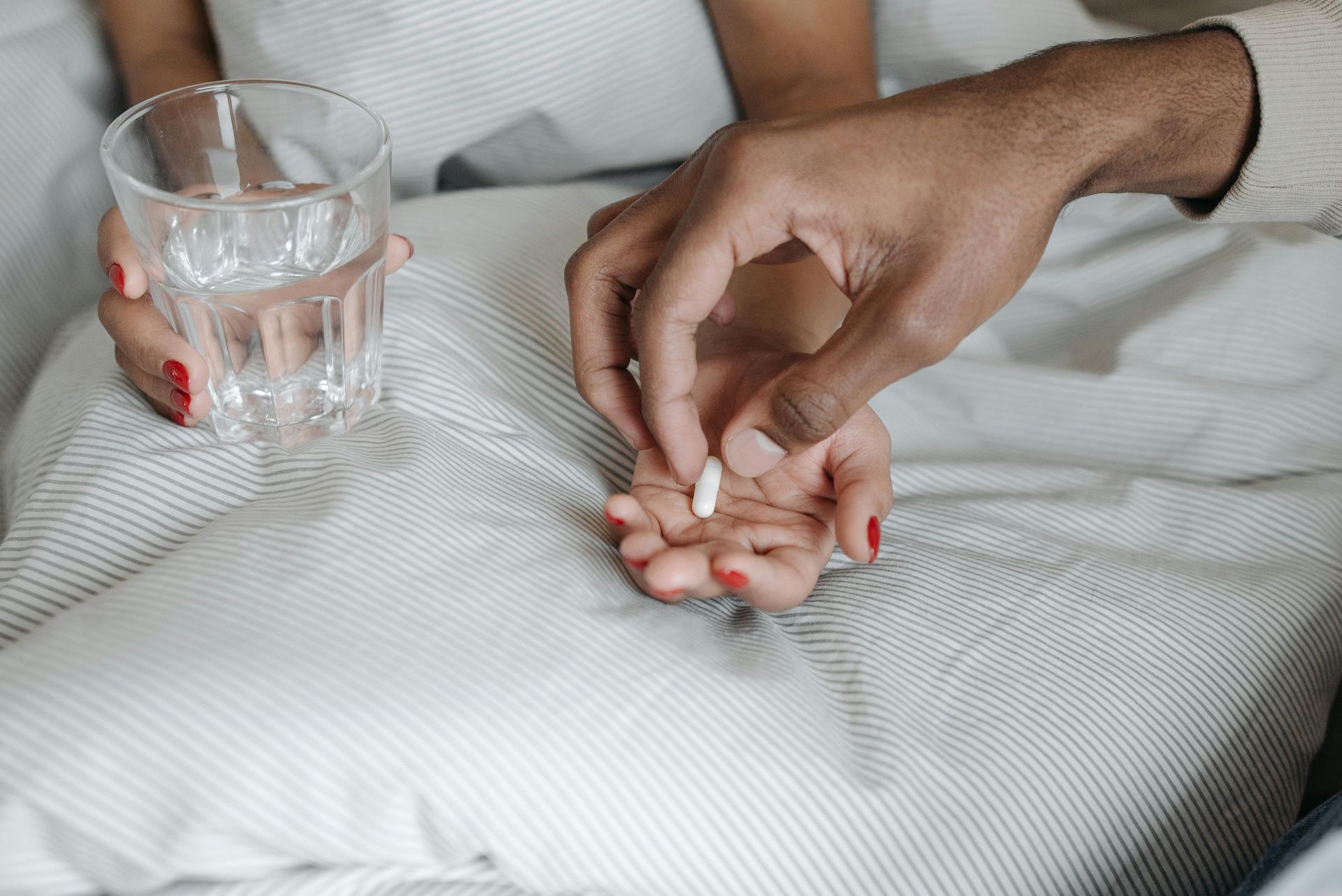 Person in bed taking pill with help from another, holding glass of water.