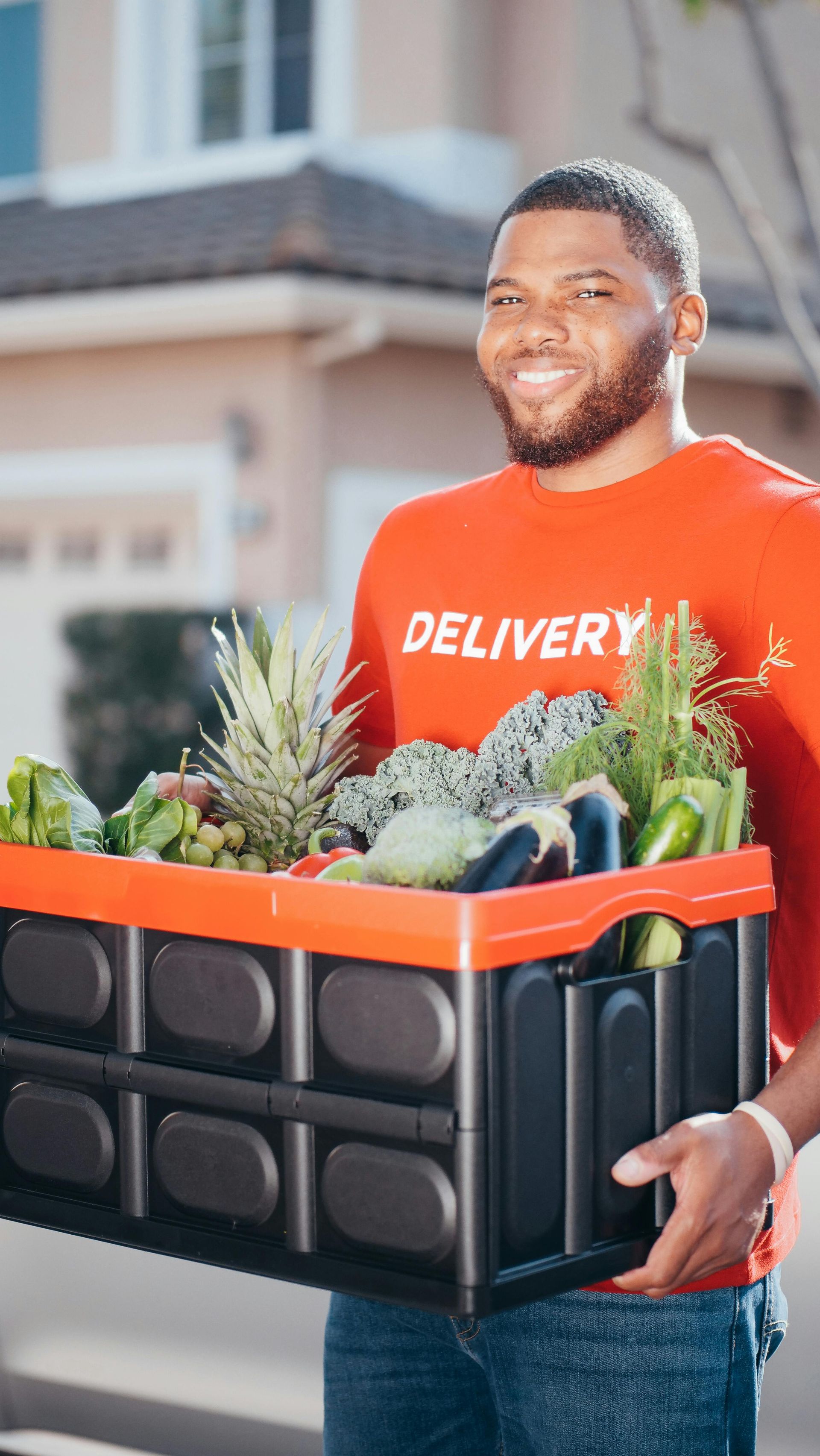 Delivery person holding a crate of groceries, smiling. Orange shirt, red