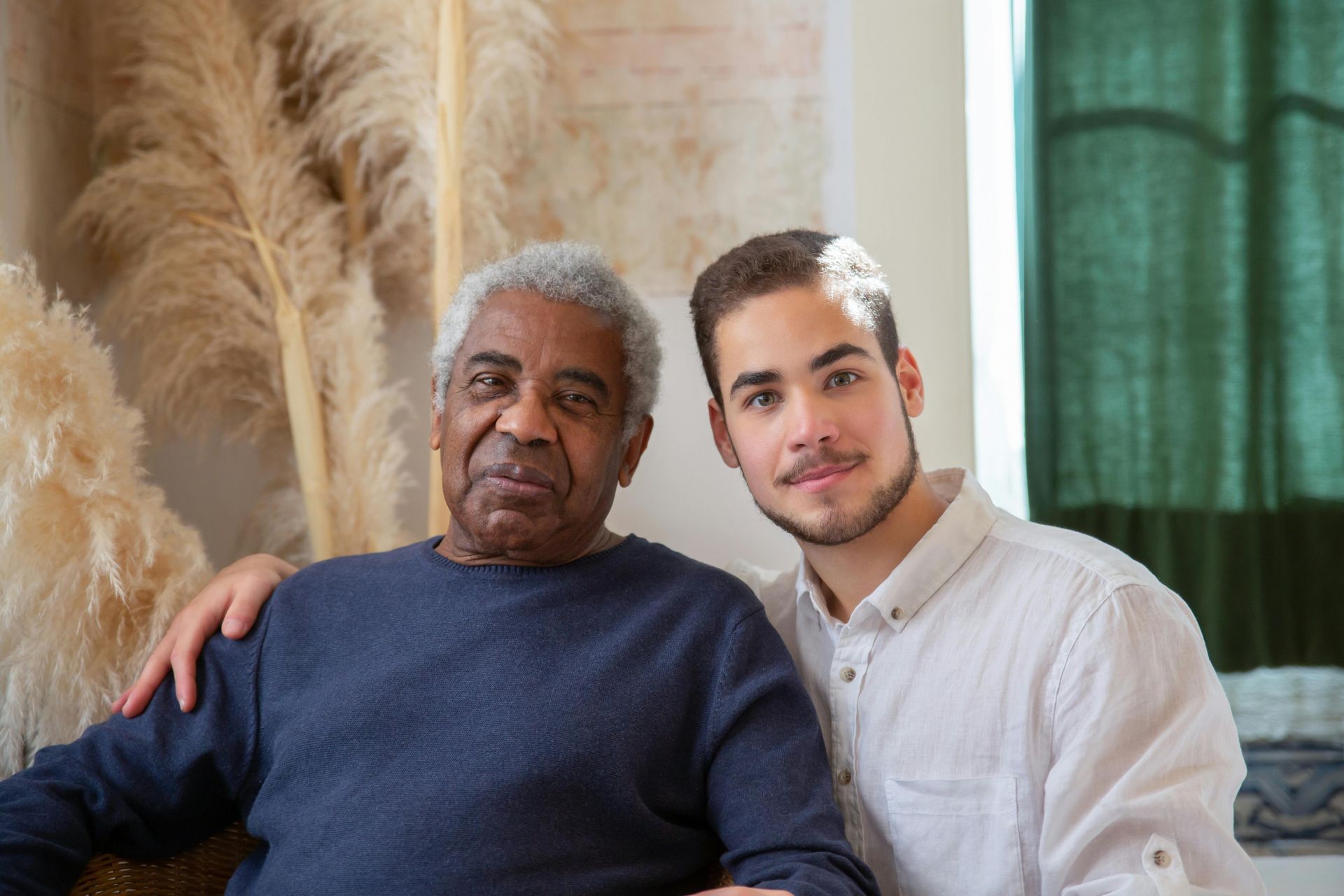 An older man in a blue sweater sits with a younger man in a white shirt, both smiling.