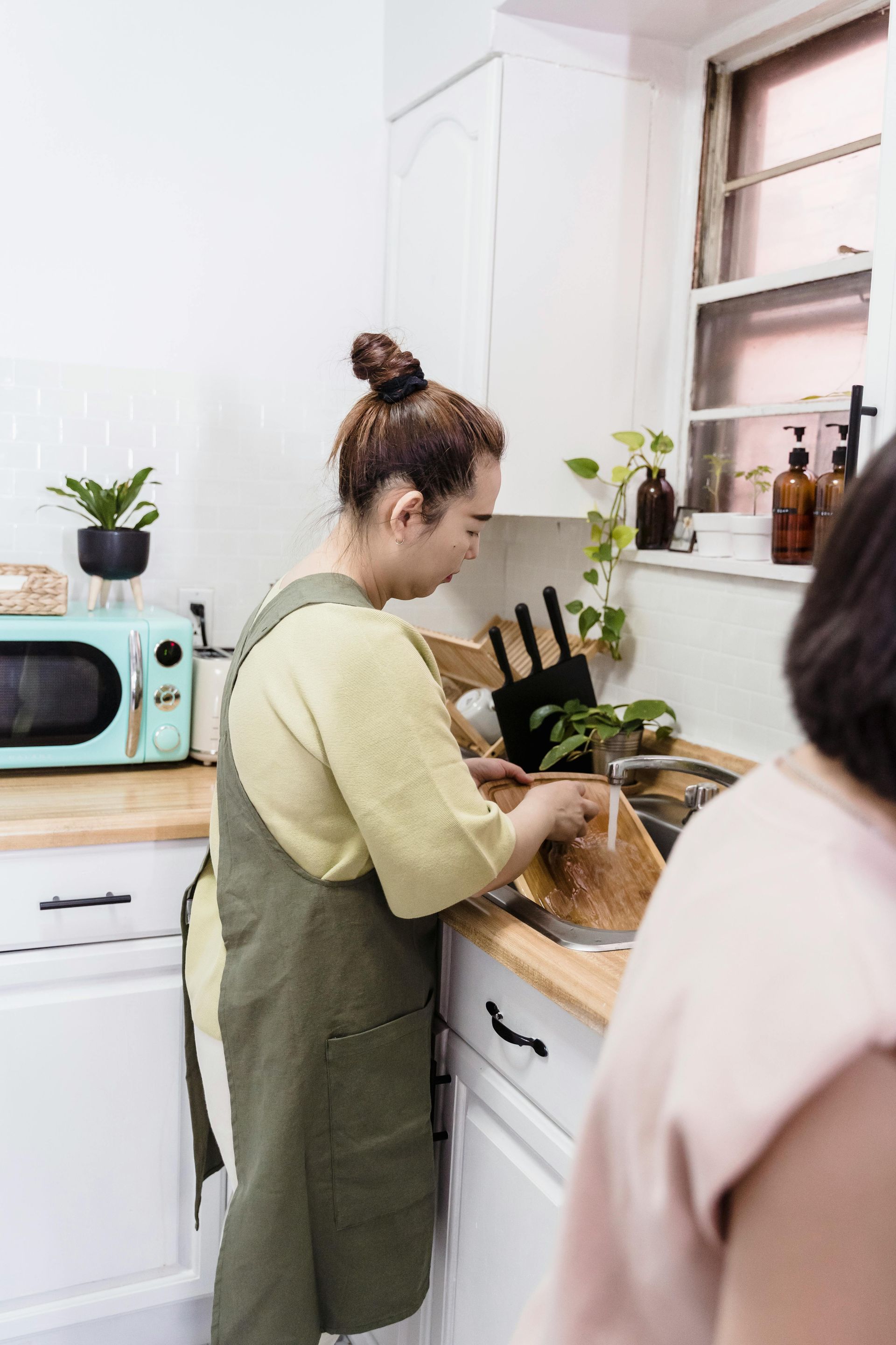 Woman washing dishes in a bright kitchen, wearing an apron, with a second person nearby.