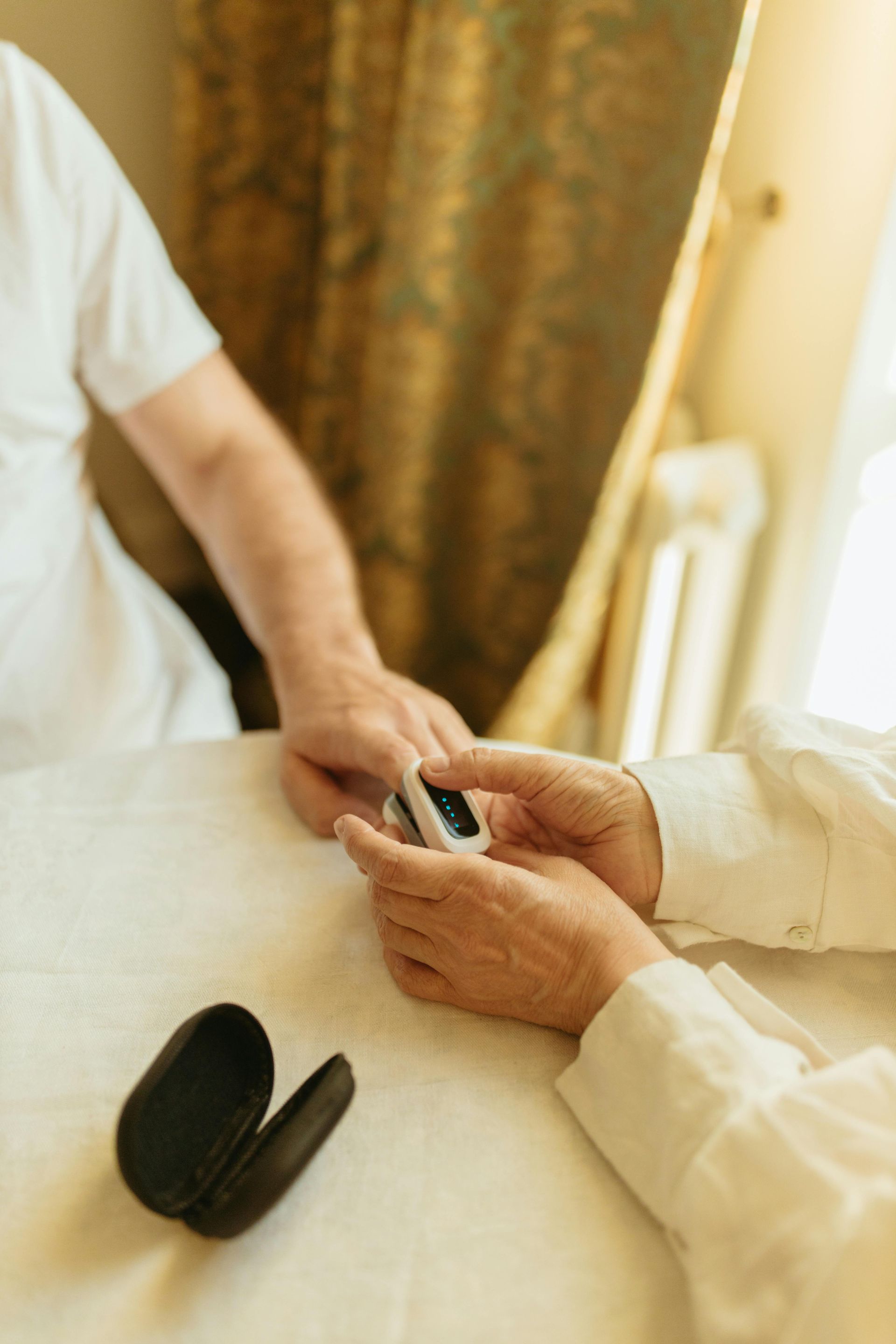 Hands using a pulse oximeter on a person's finger, indoors near a window.