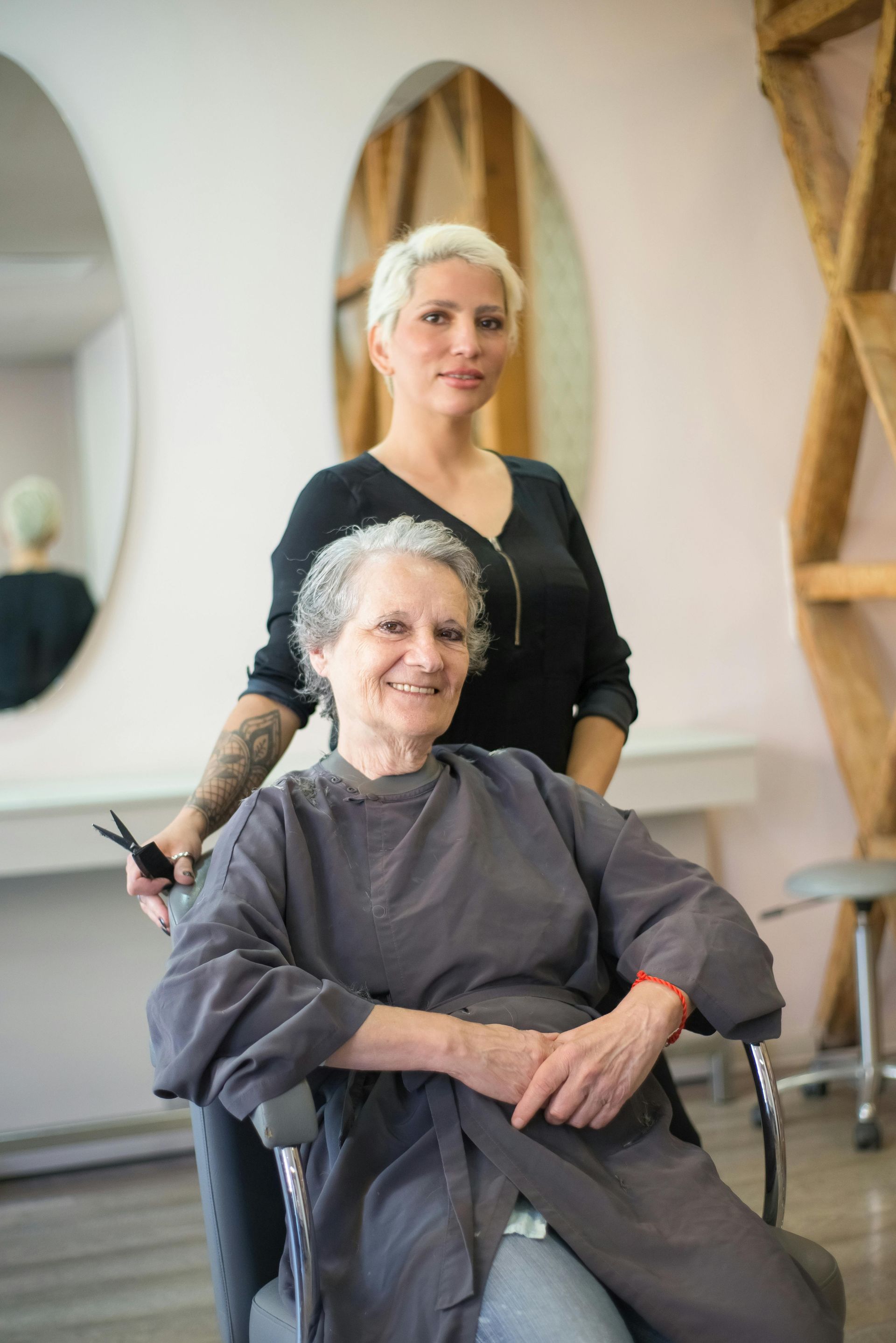 Woman with short blonde hair stands behind an older person in a salon chair, smiling.