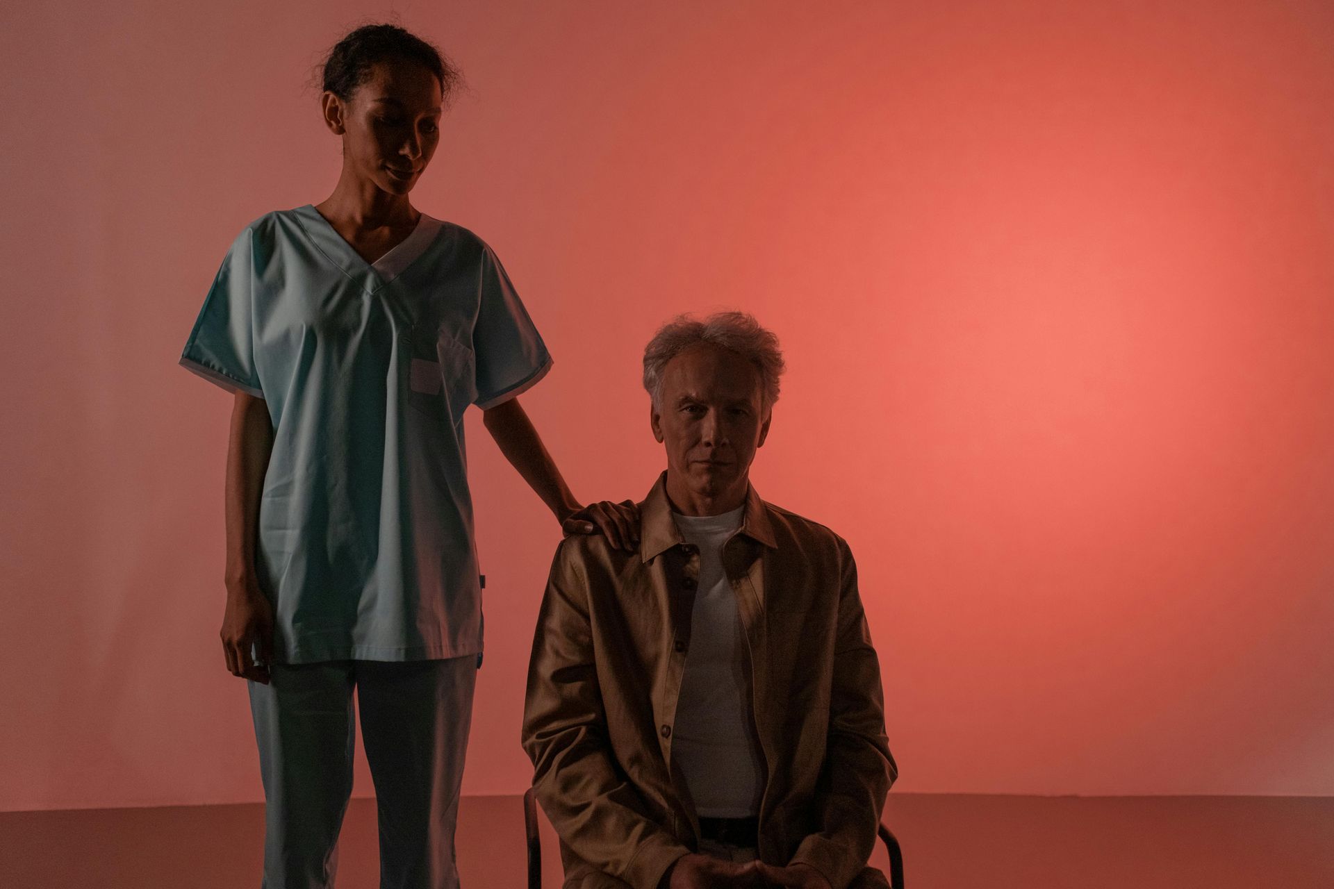 A person in a brown coat sits with a person in blue scrubs. One hand rests on the sitter’s shoulder, red backdrop.