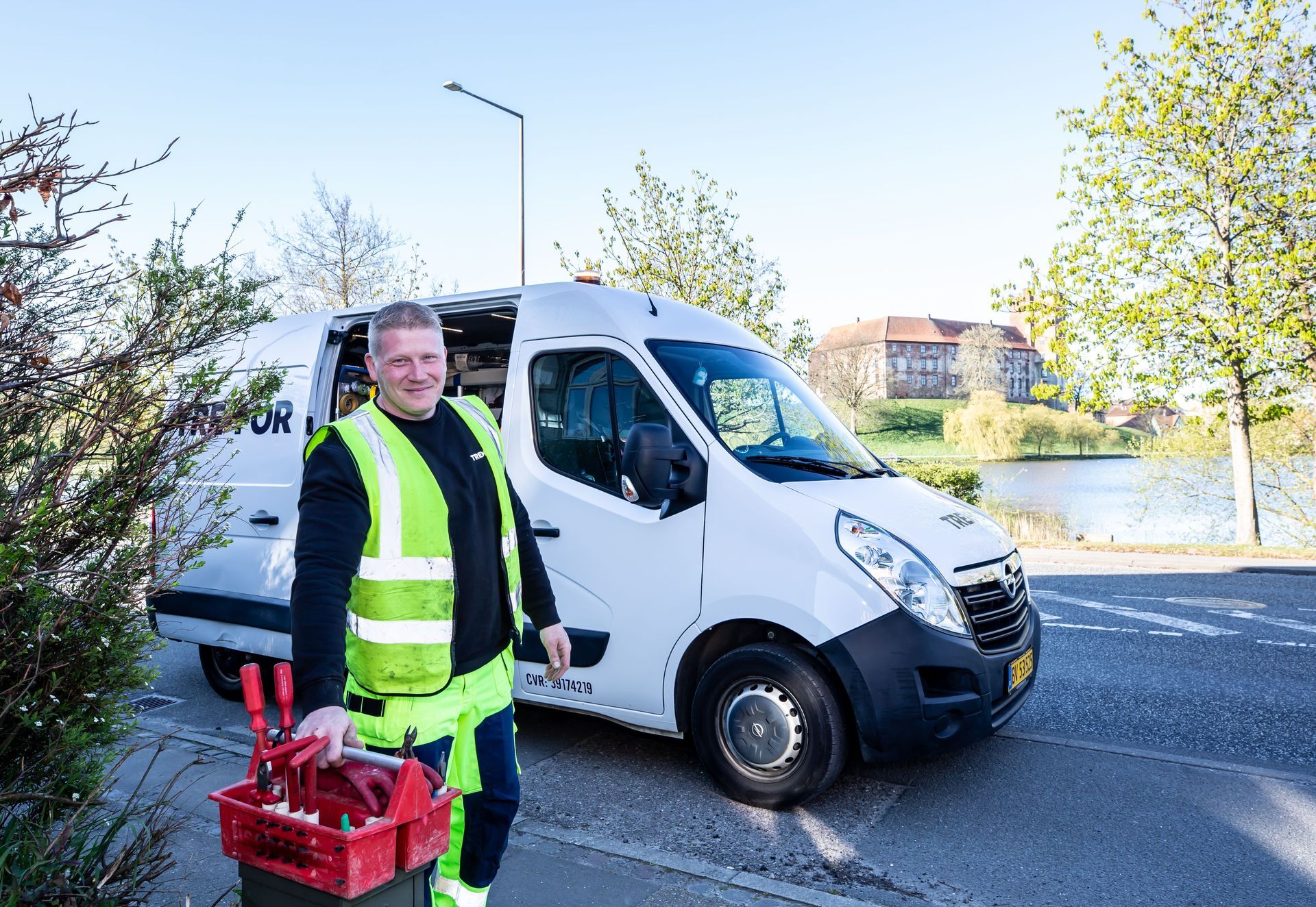Worker in neon vest holding a red crate beside a white van on a roadside.