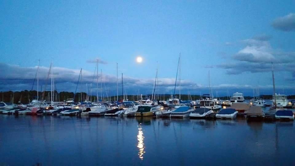 A row of boats are docked in a harbor at night with a full moon in the background.