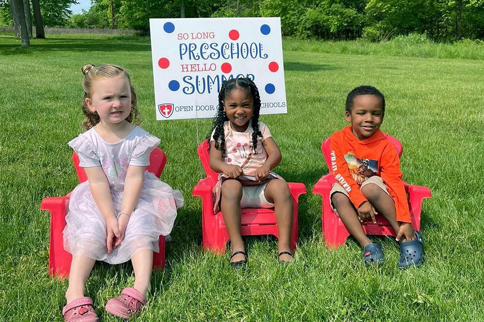 three children sit in red chairs in front of a sign that says preschool hello summer