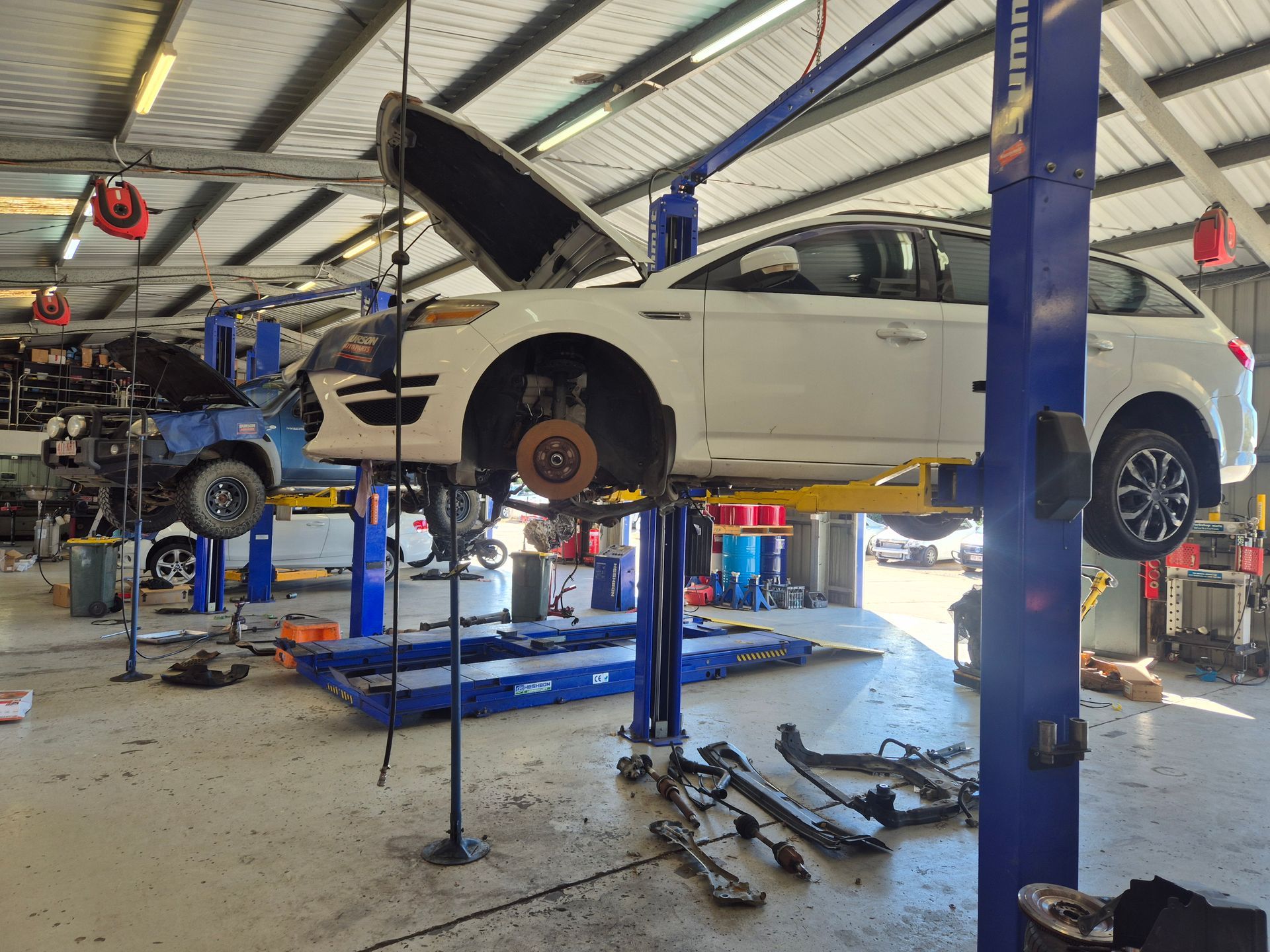 White SUV lifted on a blue hydraulic hoist in an auto repair shop, with tools and a nearby car. — Emu Park Automotive and Tyre Service in Zilzie, QLD