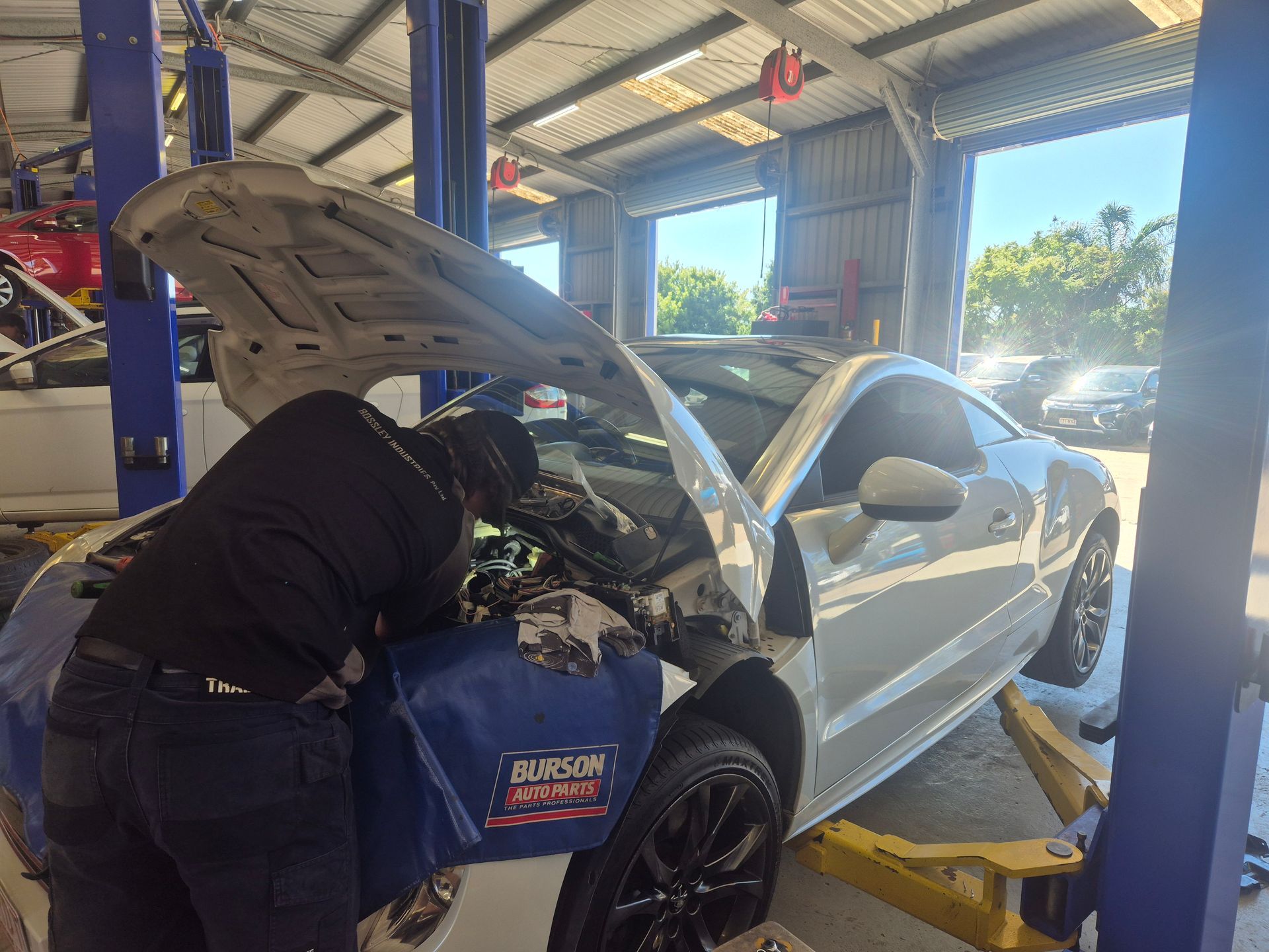 Mechanic working on a white car with open hood in a repair shop under bright daylight  — Emu Park Automotive and Tyre Service in Kinka Beach, QLD