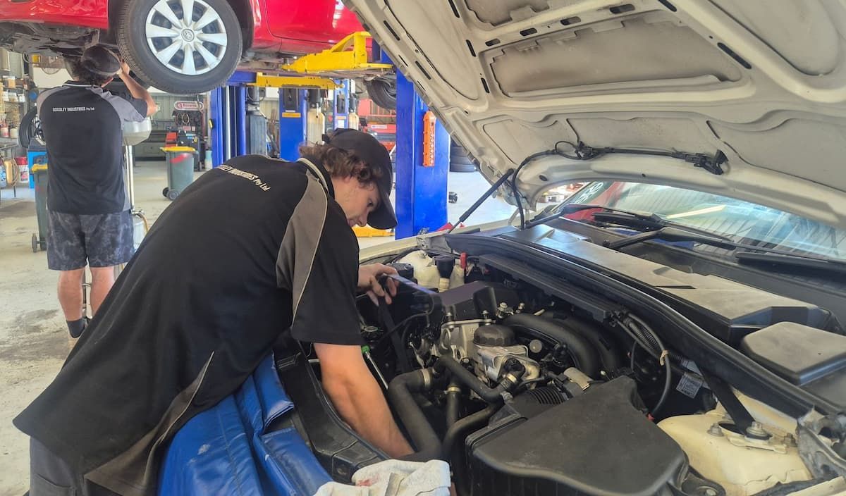 Mechanic inspecting engine bay of a blue car in an auto repair shop, with another car lifted nearby — Emu Park Automotive and Tyre Service in Zilzie, QLD
