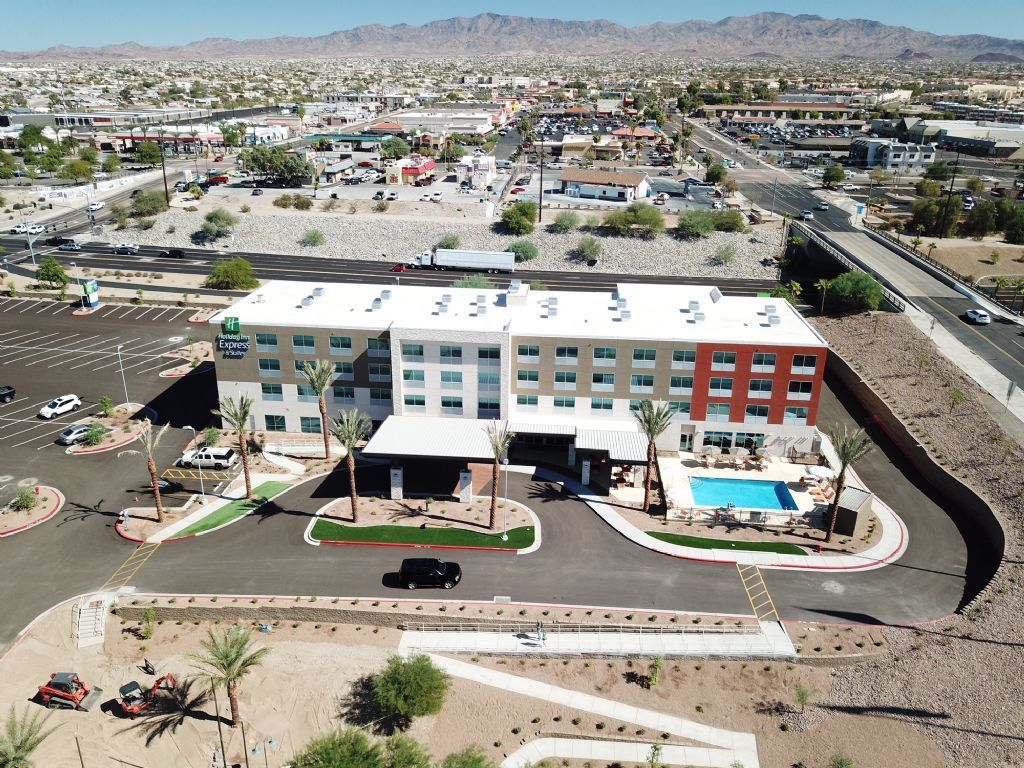 Aerial view of a four-story hotel with a pool and parking lot, set against a town and mountains.