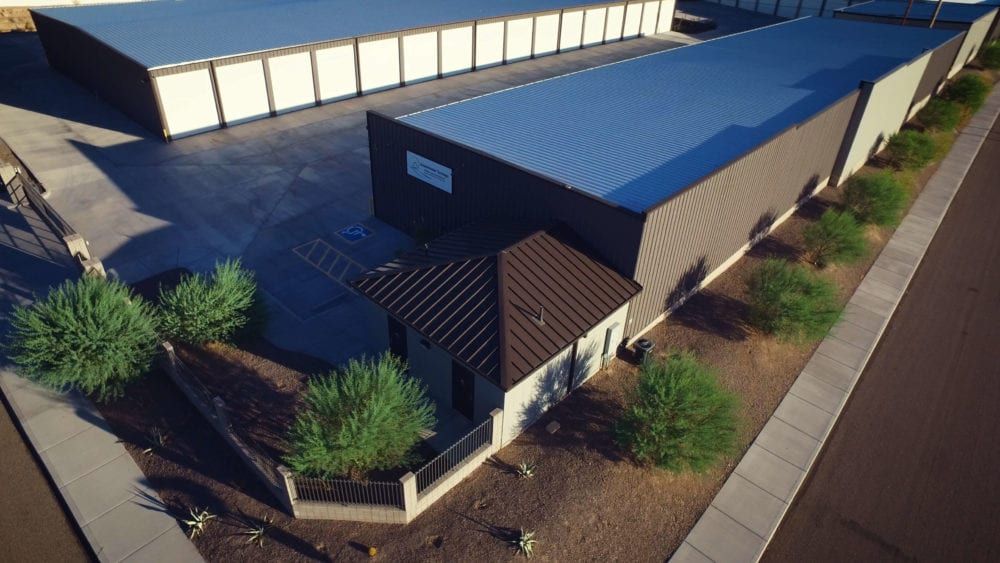 Aerial view of storage facility buildings with blue and brown roofs, shrubs, and a sidewalk.
