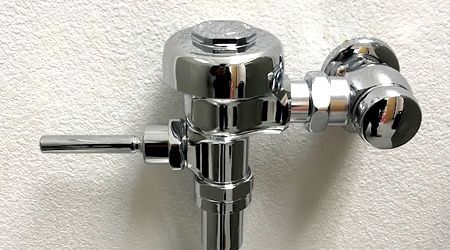 A close up of a chrome toilet flush valve on a white wall.