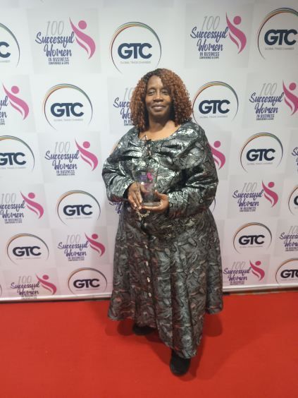 Woman in a patterned dress holding an award, standing on a red carpet in front of a backdrop with logos.
