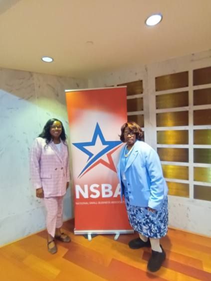 Two women stand beside an NSBA banner; one in a pink suit, the other in a blue jacket and floral skirt.