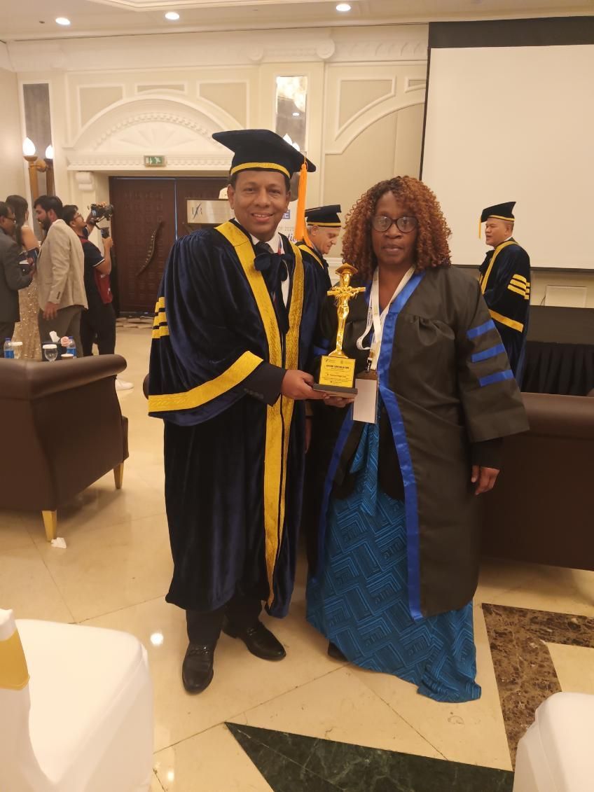 A person in academic regalia presents a trophy to a woman, indoor setting with other people.