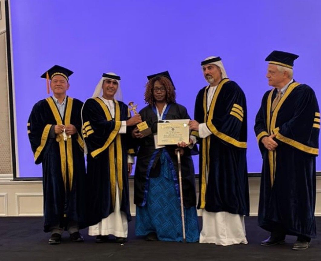 A person in graduation attire receives a diploma from officials. Blue backdrop.
