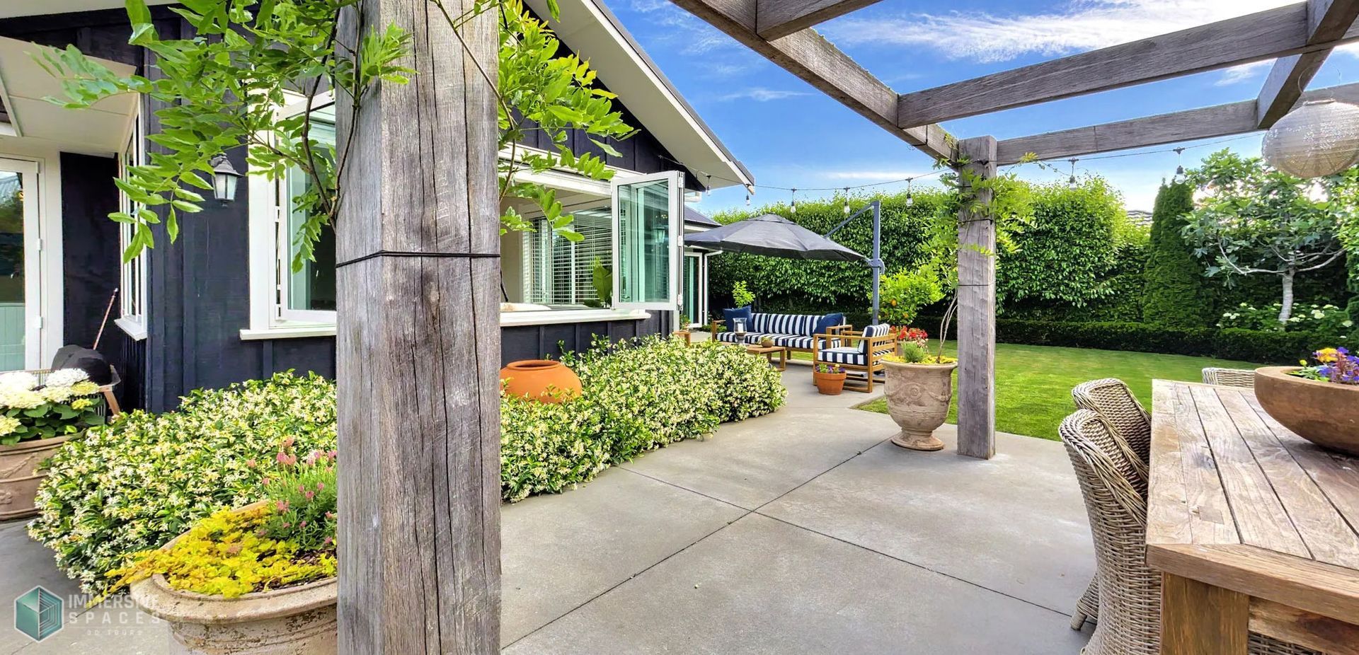 Patio with a pergola, house, and lush green garden in the background on a sunny day.
