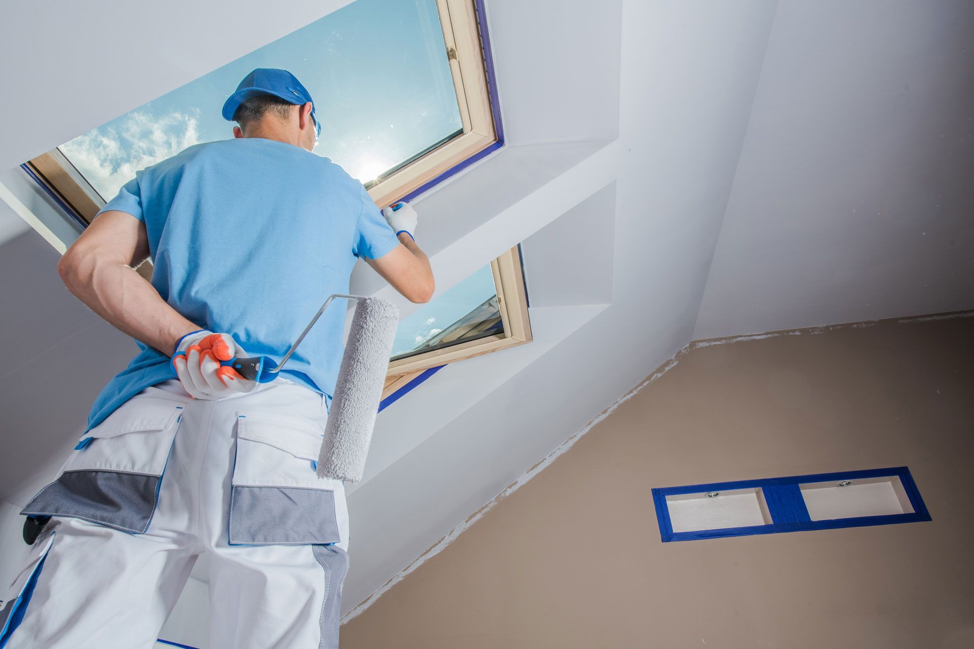 A painter in a blue shirt and white pants holds a paint roller while working on a slanted ceiling near two skylights.