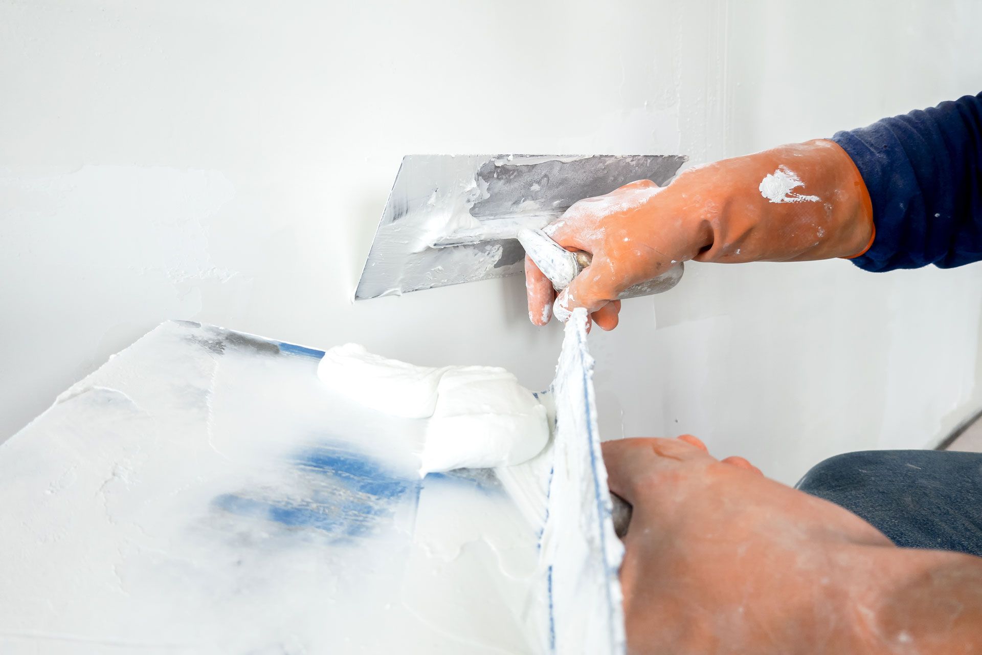 A person wearing gloves uses a metal trowel to smooth white joint compound onto a wall.