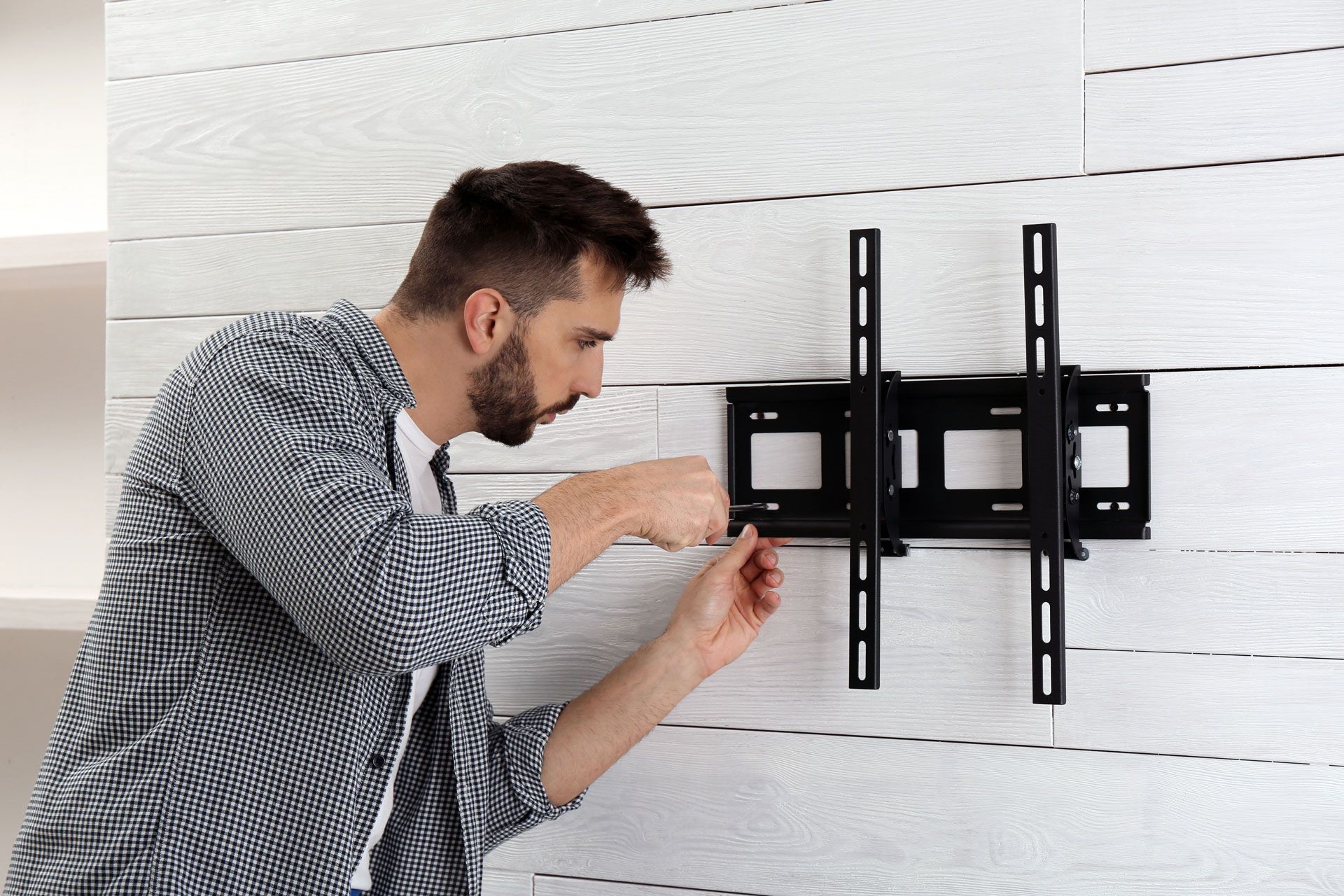 A person in a plaid shirt installs a black TV wall mount on a white wood-paneled wall.