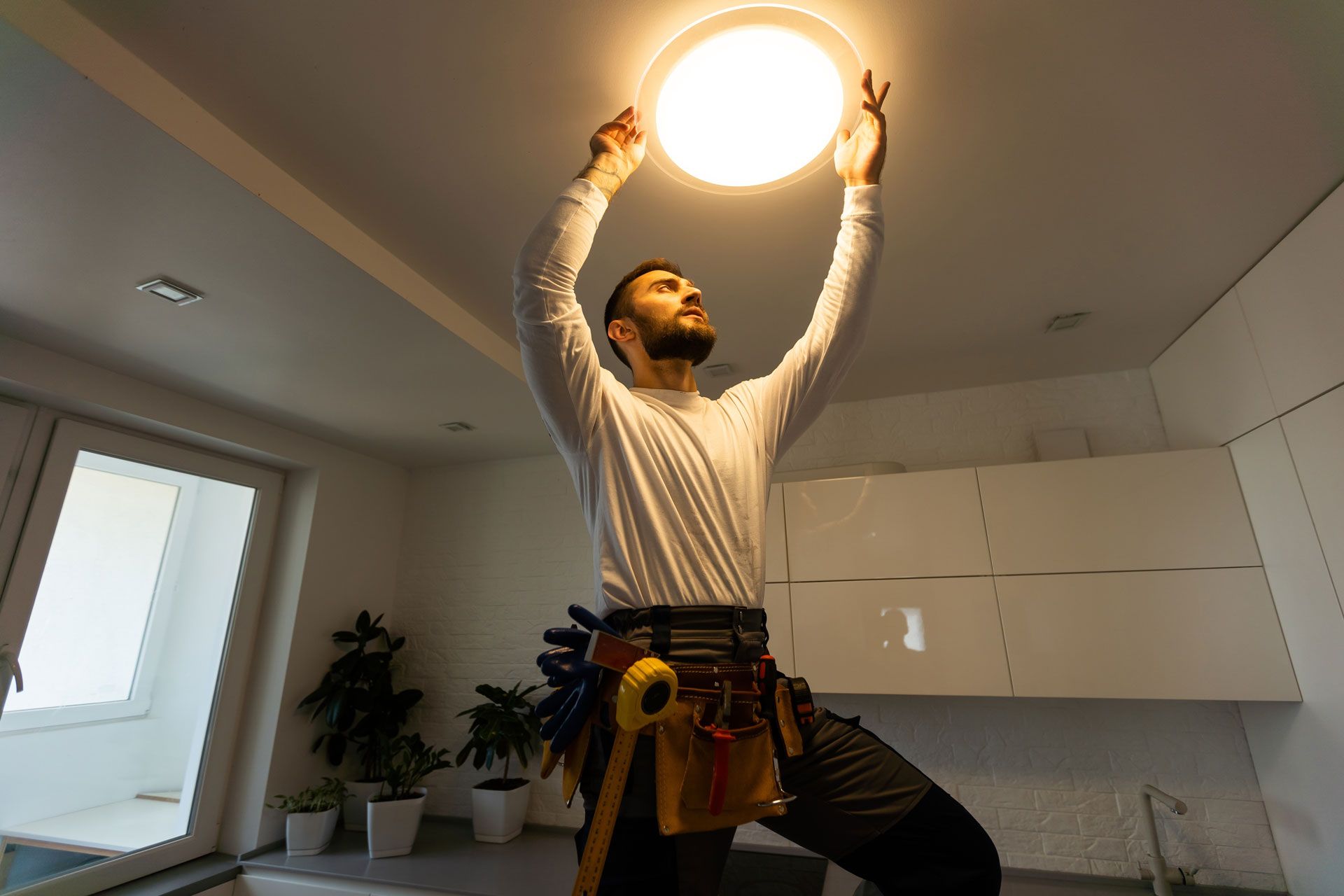 A person in a tool belt installs a circular ceiling light fixture in a modern kitchen.