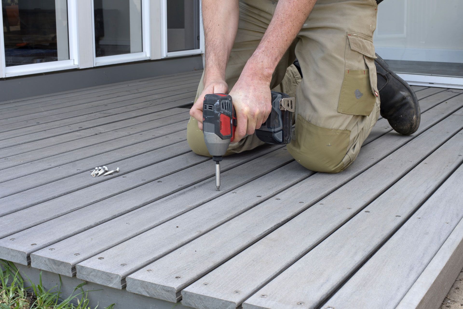 A person kneels on a grey deck, using a power drill to drive a screw into the wood surface.
