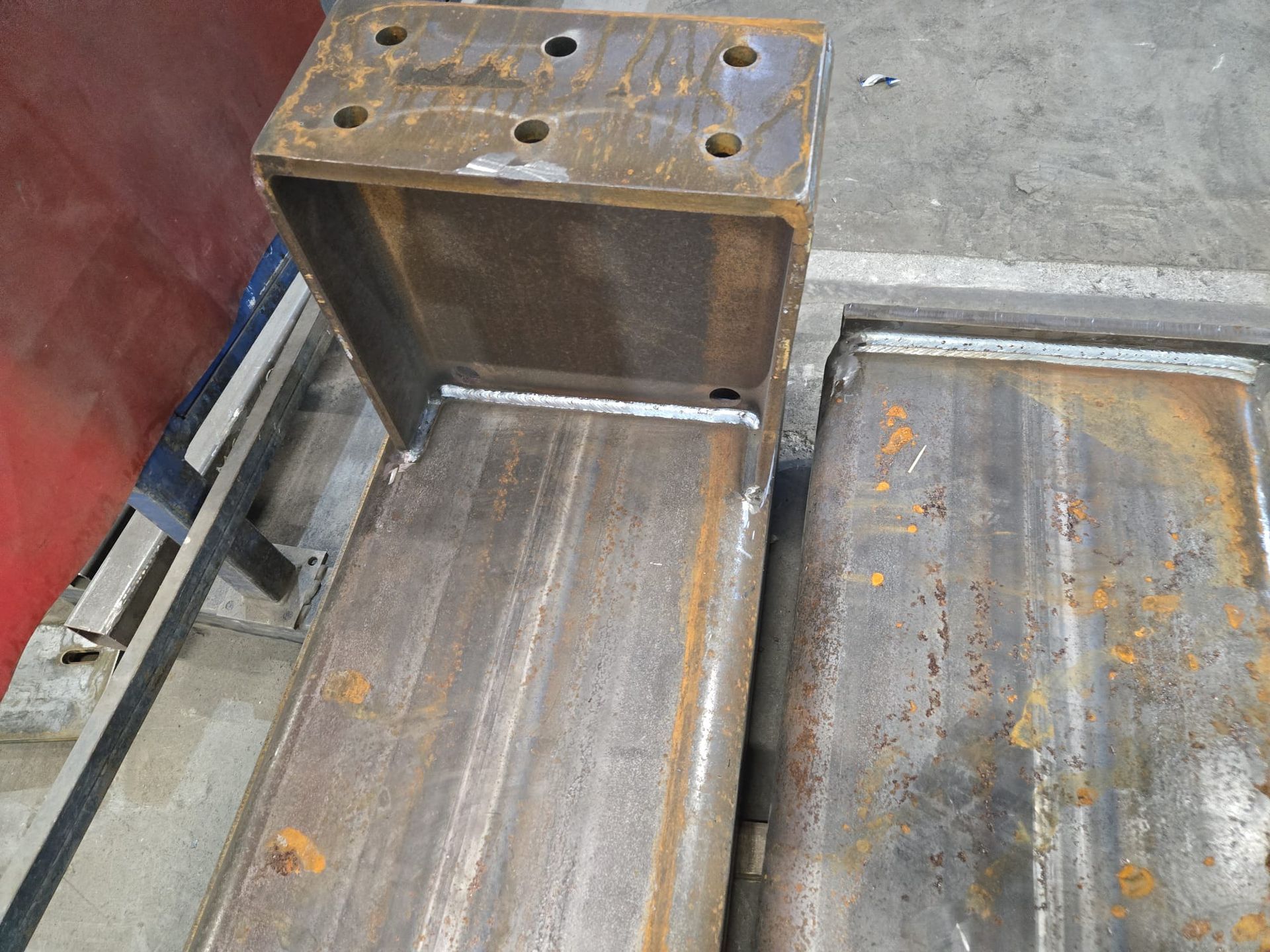 Primo piano di una trave metallica marrone non rifinita, con una staffa saldata e diversi fori circolari per bulloni.