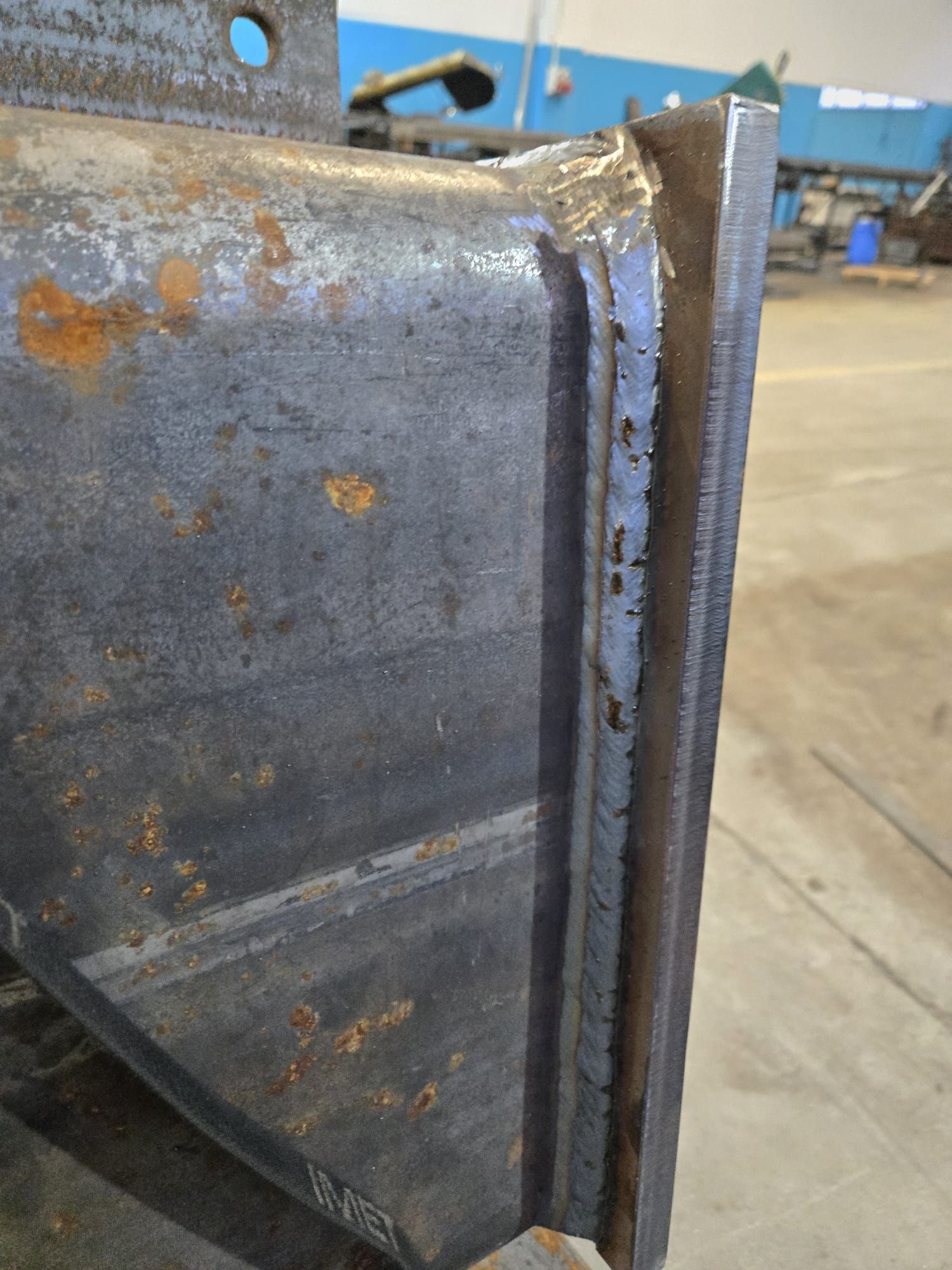 Primo piano di una giunzione in acciaio strutturale con una spessa saldatura verticale che unisce due piastre metalliche in un ambiente di officina.