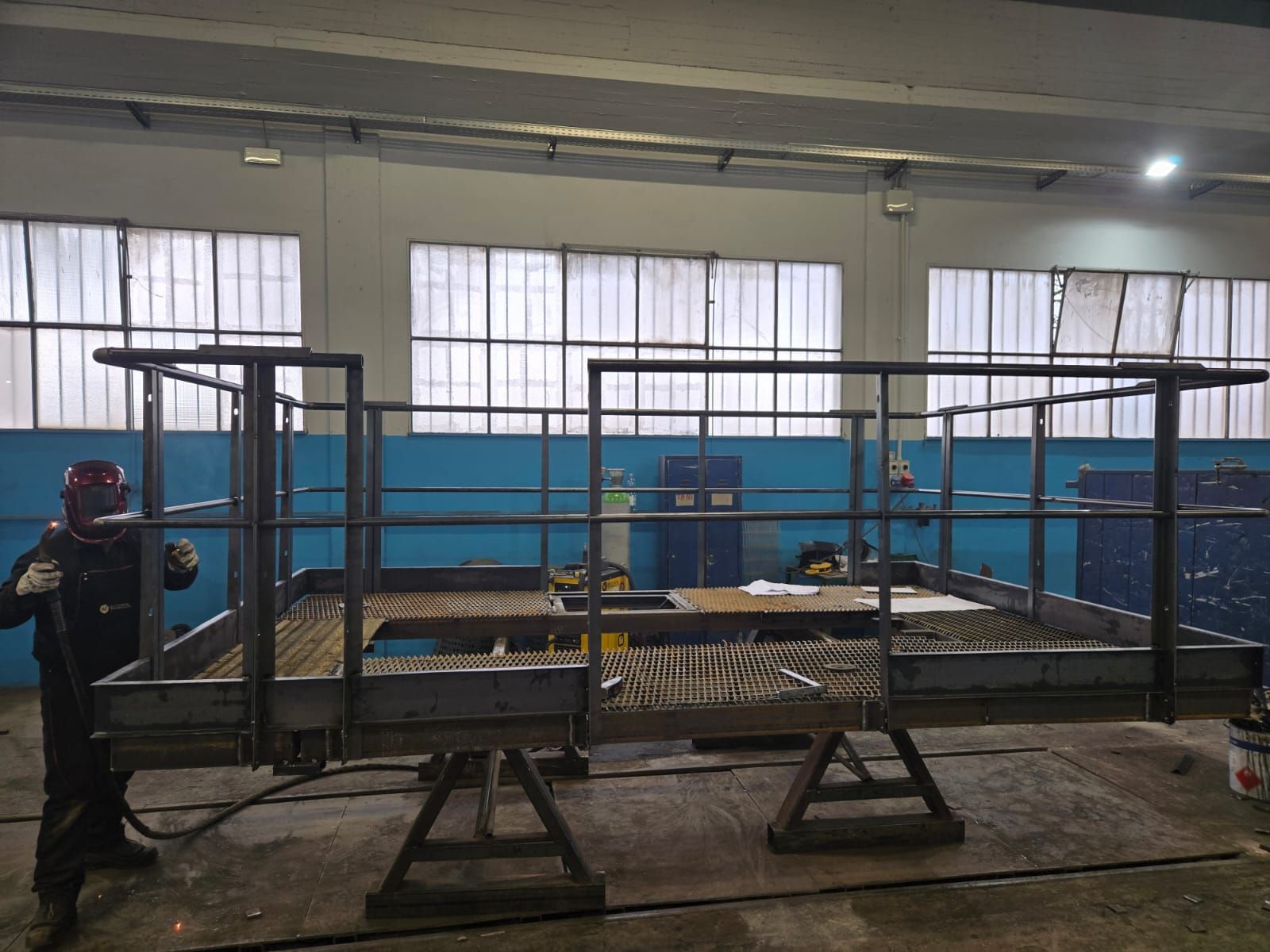 Un operaio con indumenti protettivi salda una struttura metallica a piattaforma all'interno di un'officina industriale ben illuminata.