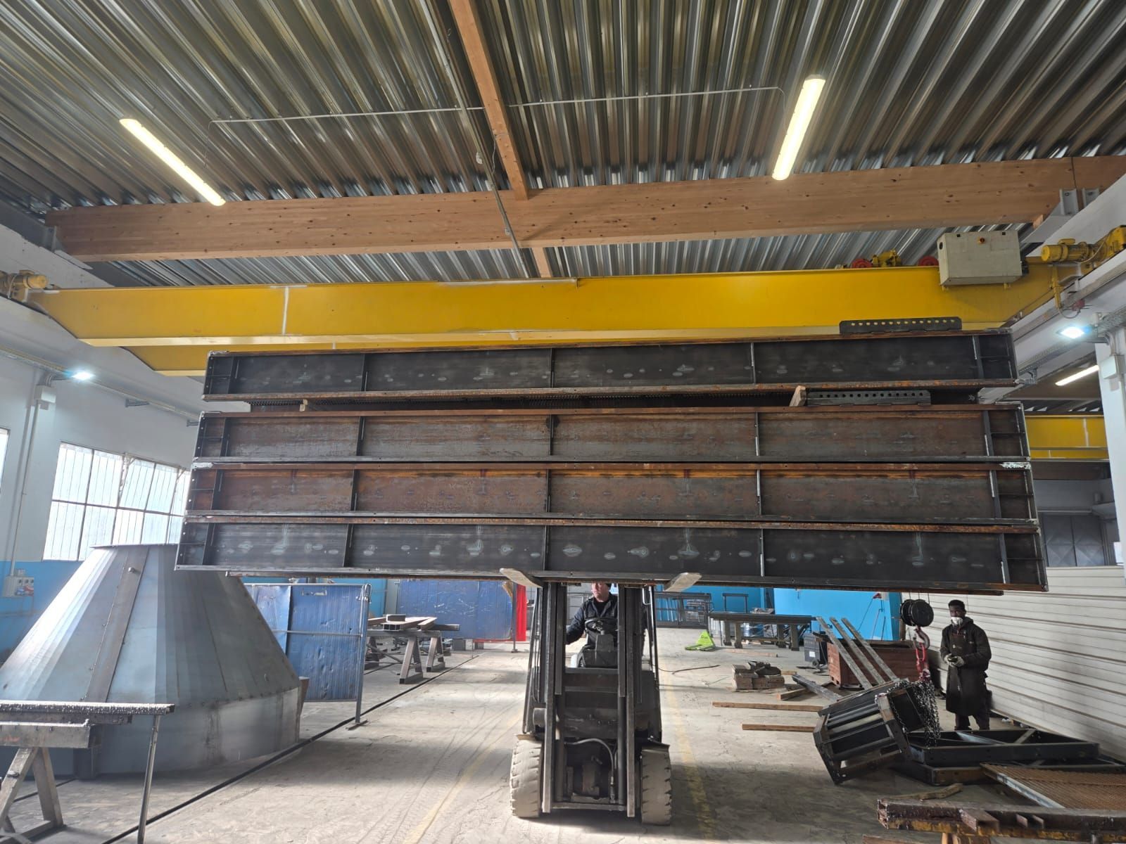 In un'officina industriale, un carrello elevatore solleva una grande pila di travi metalliche scure con una gru a ponte gialla.