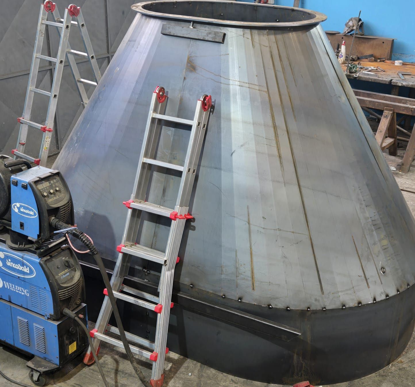 Una grande struttura conica in metallo all'interno di un'officina, con due scale appoggiate ad essa e una saldatrice blu nelle vicinanze.
