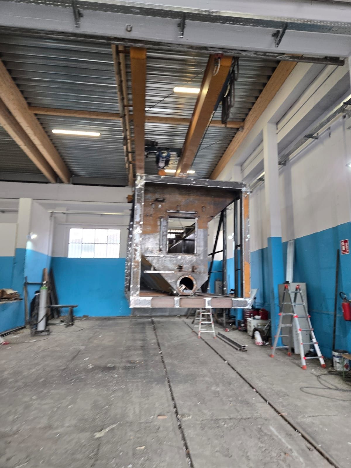 In una fabbrica con pareti dipinte di blu, una grande struttura metallica industriale è sospesa a una gru a ponte.