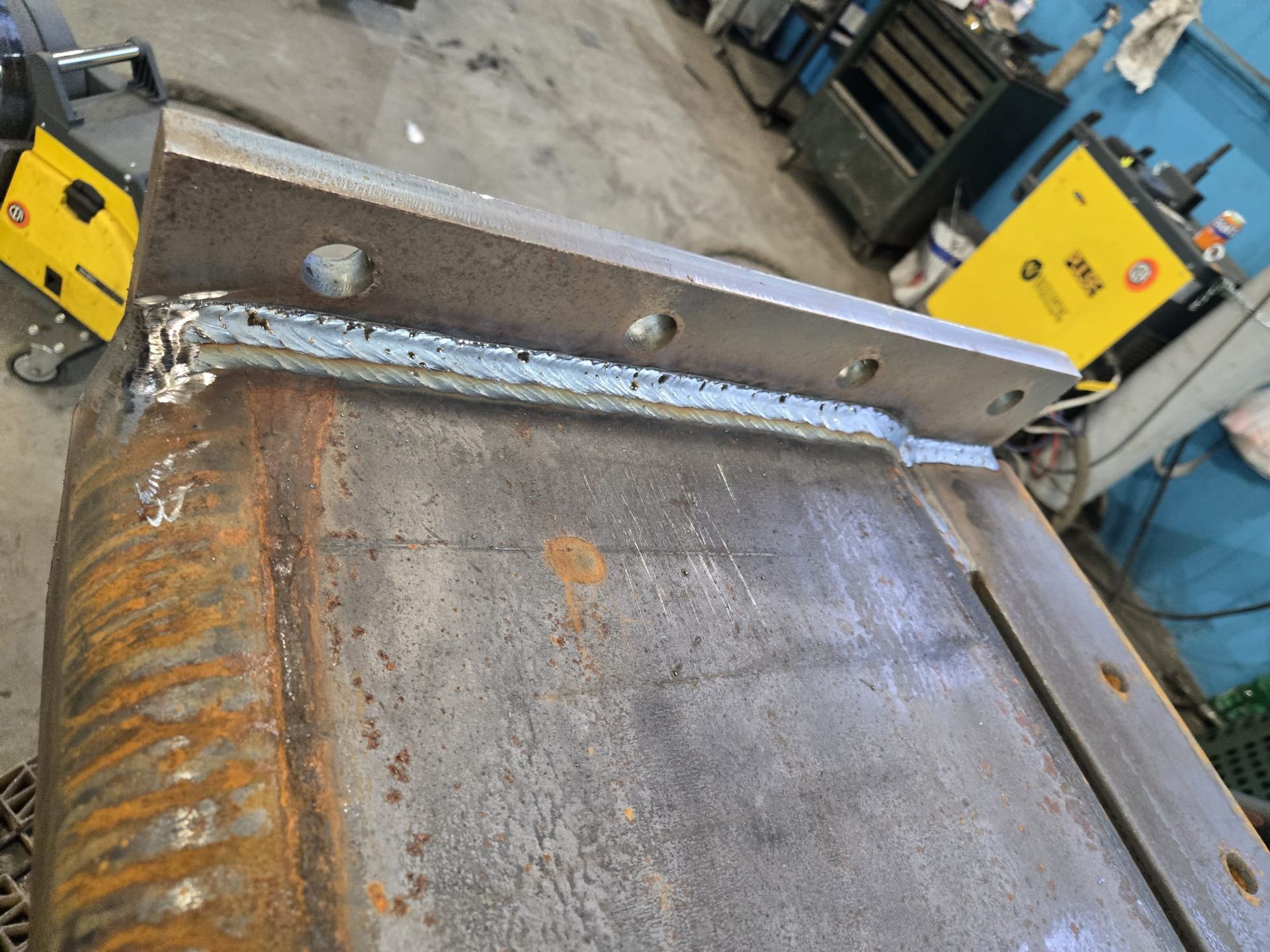 In un'officina, un cordone di saldatura fresco e di colore chiaro unisce una spessa piastra d'acciaio a una trave metallica arrugginita.