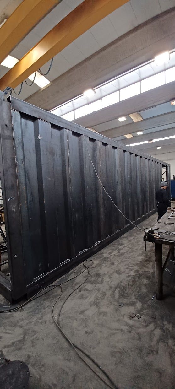 Un lungo container nero in fase di allestimento all'interno di un'ampia officina industriale illuminata da luci intense sul soffitto.