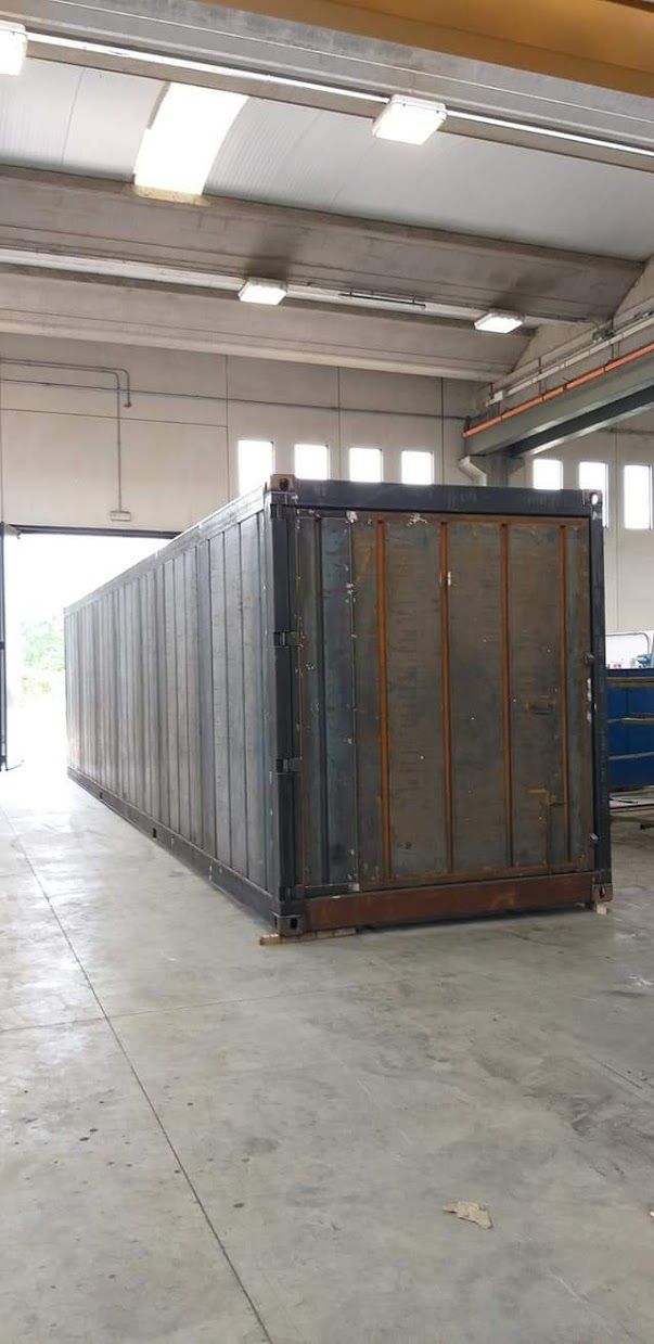 Un container per spedizioni in lamiera ondulata scura si trova all'interno di un grande magazzino industriale illuminato dall'alto.