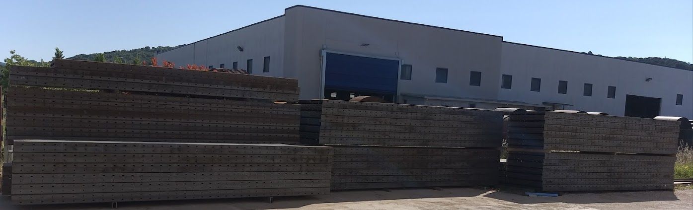 In primo piano, all'esterno di un lungo edificio bianco adibito a magazzino, sotto un cielo azzurro limpido, si stagliano travi di legno scuro impilate.