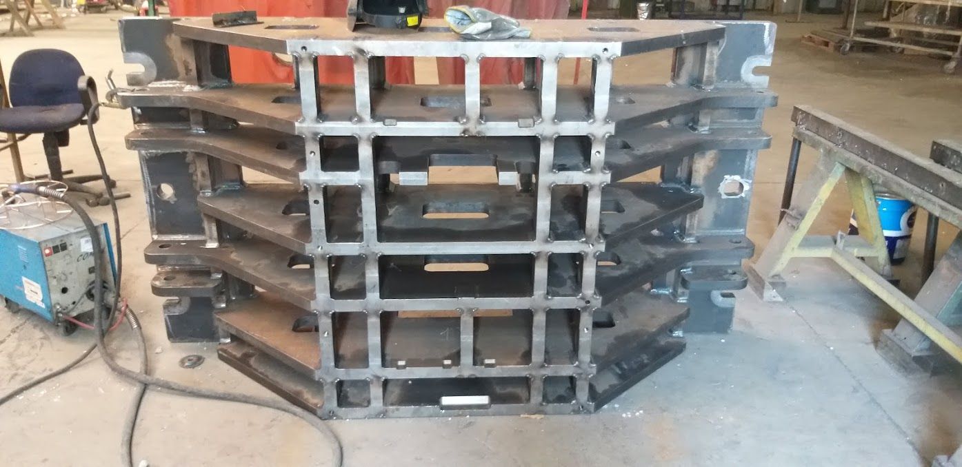 Un'officina di lavorazione dei metalli che mostra una grande struttura reticolare rettangolare in acciaio appoggiata su un robusto telaio industriale.