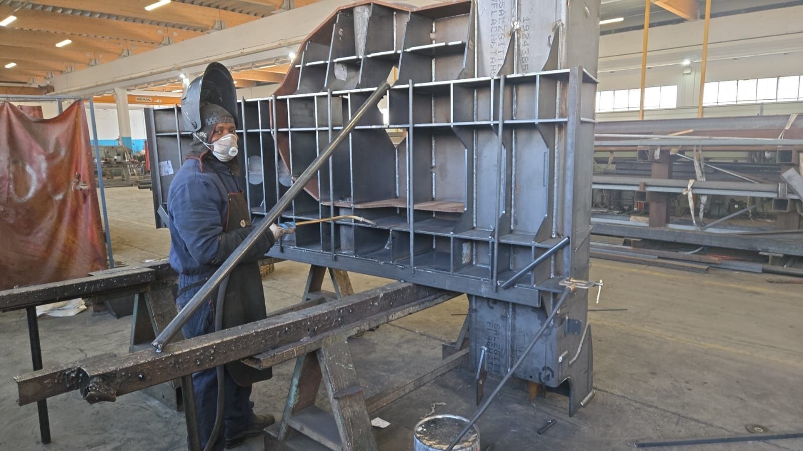Un saldatore con indumenti protettivi lavora su un componente strutturale metallico di grandi dimensioni e di difficile realizzazione all'interno di una fabbrica.